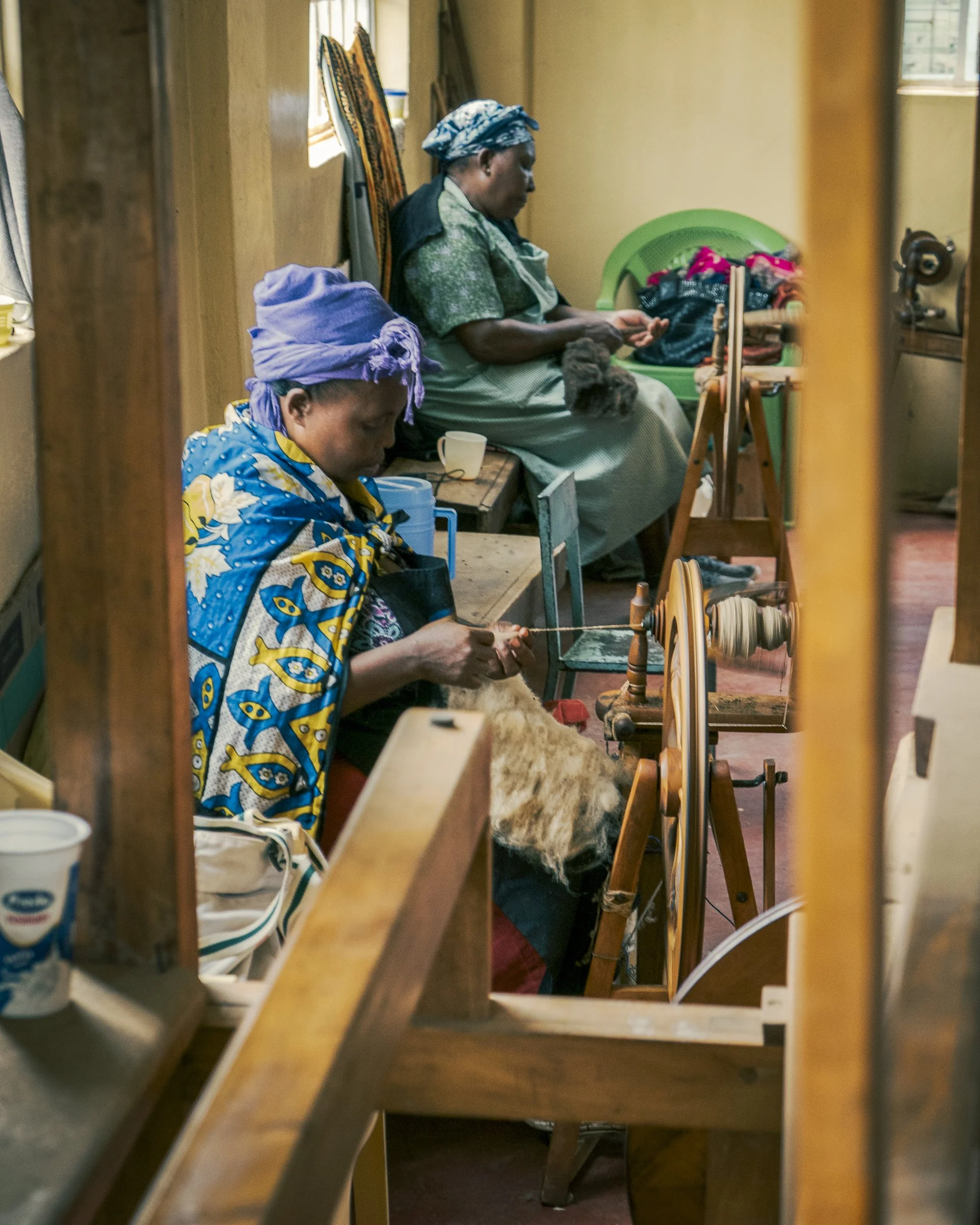 A woman and a girl are sitting in a room engaged in textile work. The woman is sewing or weaving on a loom, while the girl, dressed in a colorful patterned garment and purple headscarf, appears to be working or resting nearby. The room has sunlight c