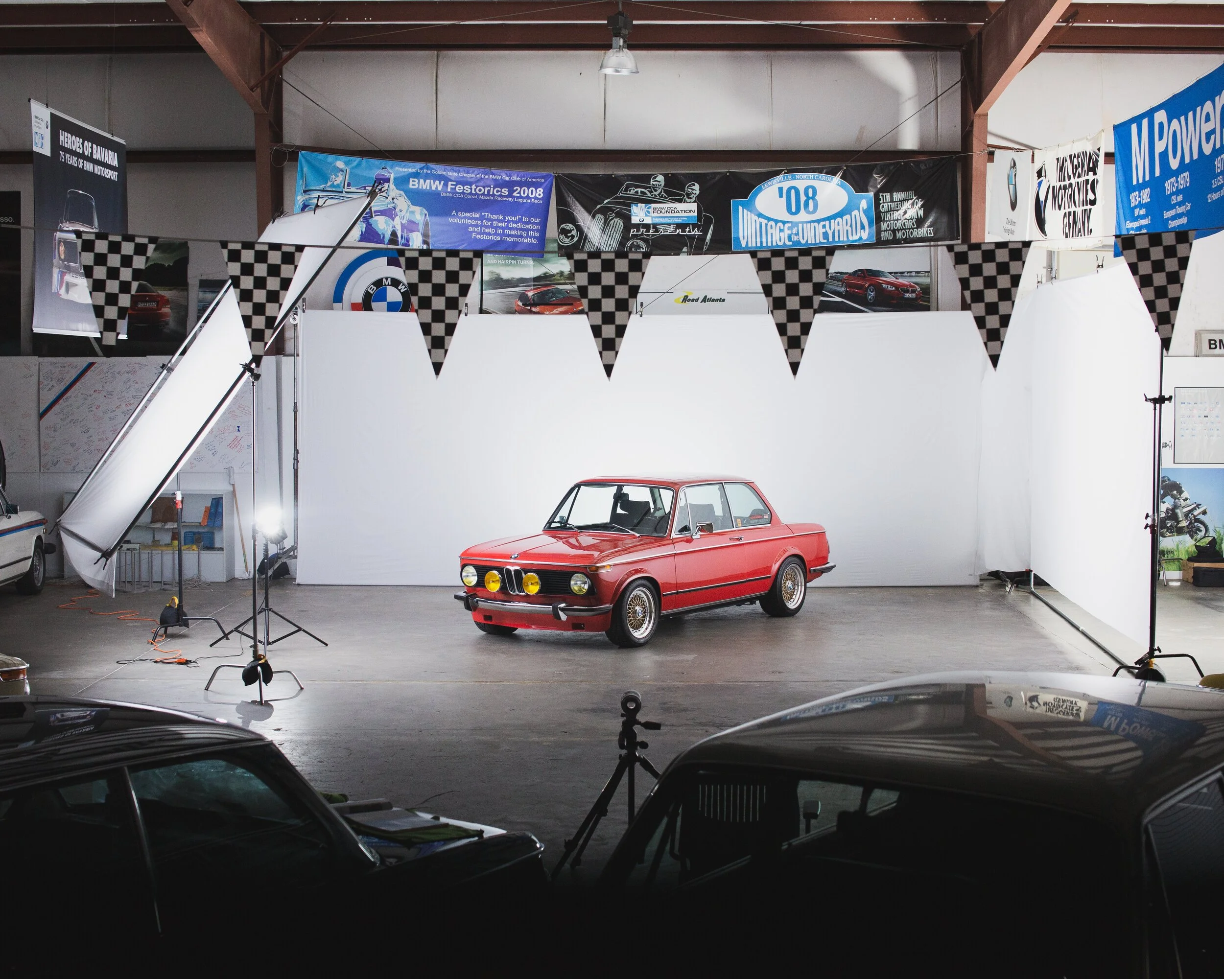 Indoor setting with a red vintage car in front of a white backdrop, surrounded by photography lighting equipment and a checkered flag banner, and additional cars partially visible in the foreground.