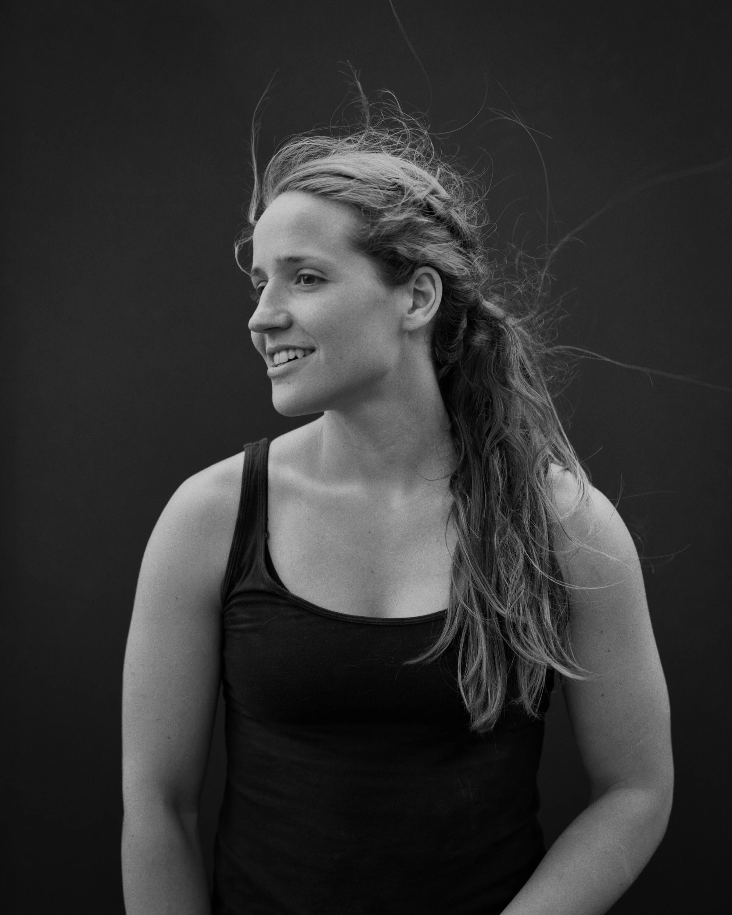 Black and white portrait of a smiling young woman with long hair, wearing a tank top, against a plain background.