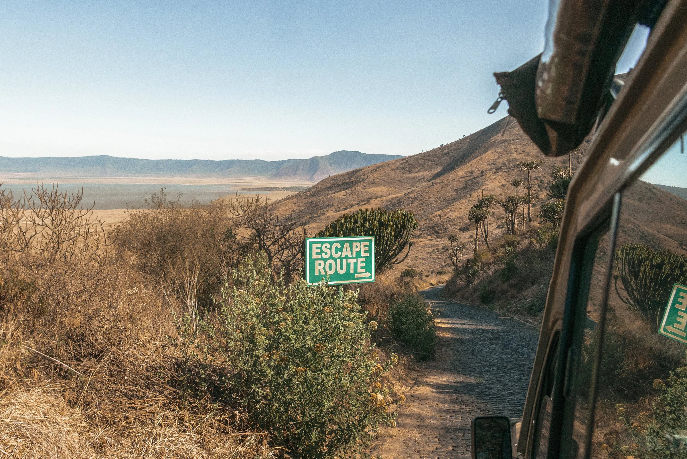 Scenic view of desert mountains and vegetation with a green sign reading 'Escape Route' and an arrow pointing right, seen from the window of a vehicle.