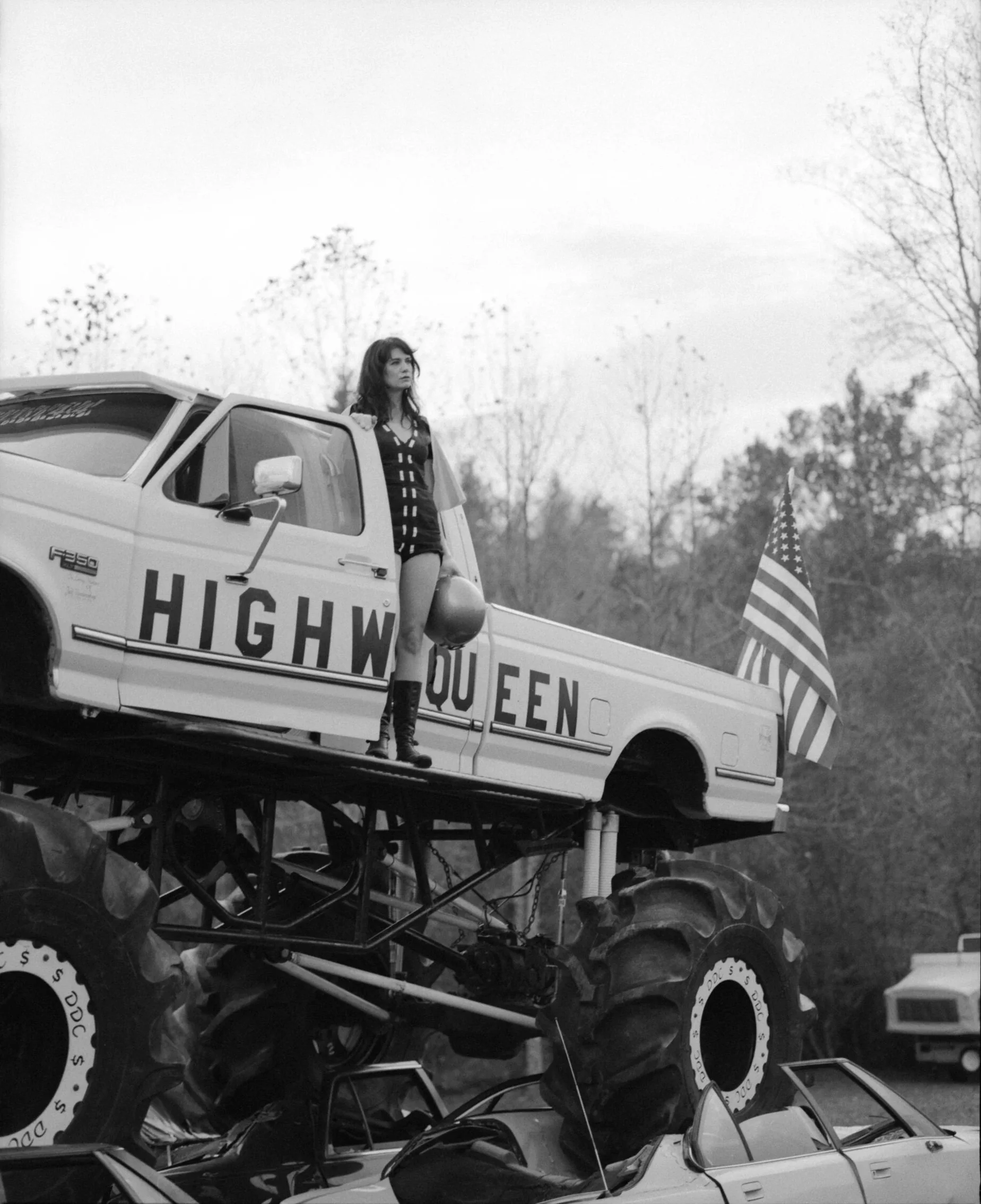 A woman standing inside a large, homemade, rugged vehicle or go-kart with large tires and a high chassis, with an American flag attached, outdoors with trees in the background.