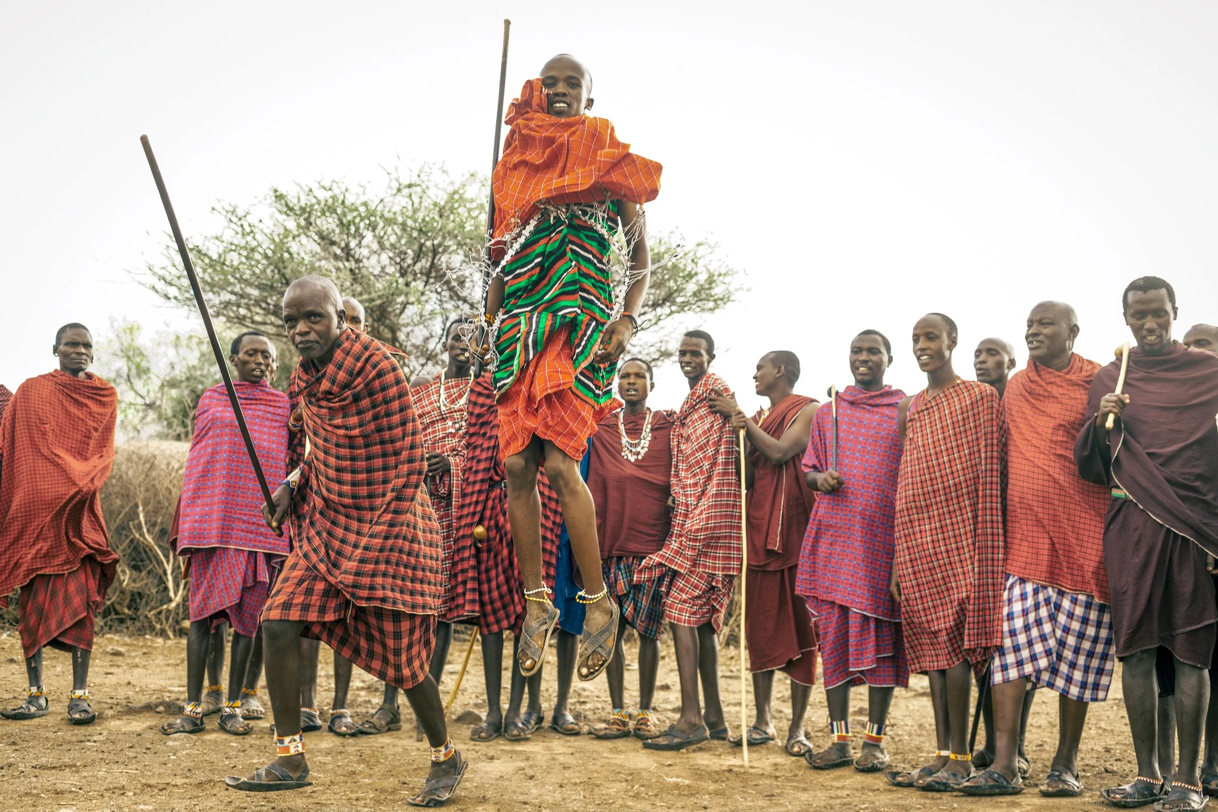 A group of Maasai warriors in traditional attire, with one jumping and smiling in the foreground. Others stand behind him with spears, wearing red, blue, and checkered shukas, in an open outdoor setting.