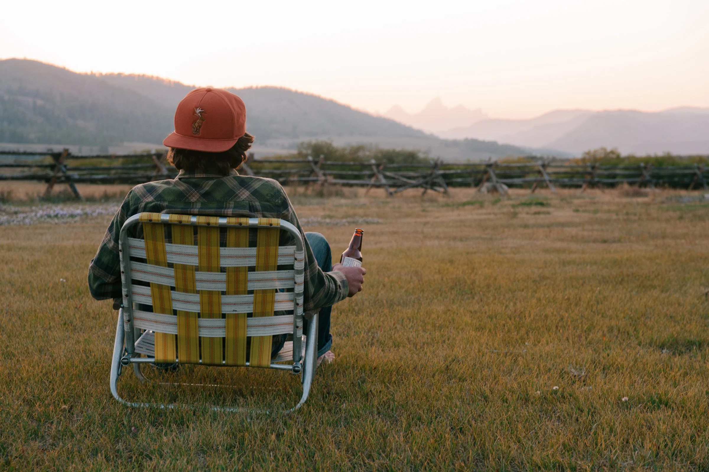Person sitting in a yellow and gray striped lawn chair, wearing a brown hat with a deer emblem, plaid shirt, and holding a bottle, facing a scenic mountain landscape during sunset.