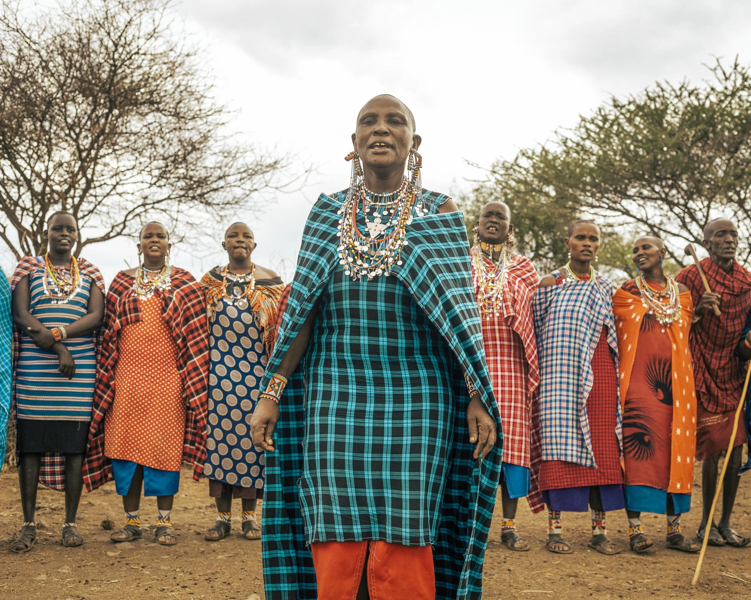 A group of Maasai women dressed in colorful traditional attire, jewelry, and beaded accessories, standing outdoors with trees and a cloudy sky in the background. The woman in the foreground wears a blue and black checked shuka, while others wear simi