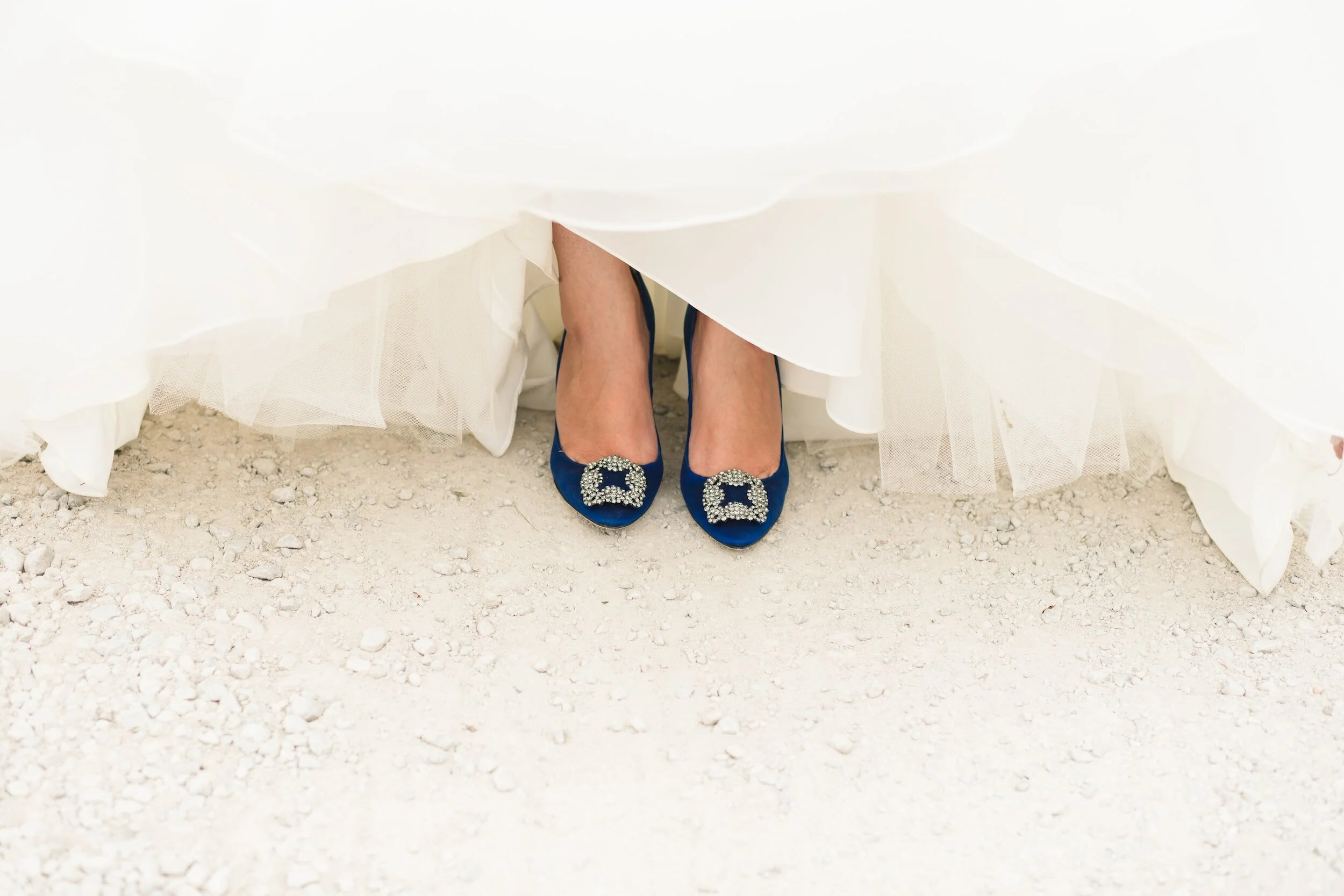 Close-up of a bride's out-of-focus legs and feet, wearing blue heels with rhinestone embellishments, beneath her white wedding dress, standing on a gravel surface.