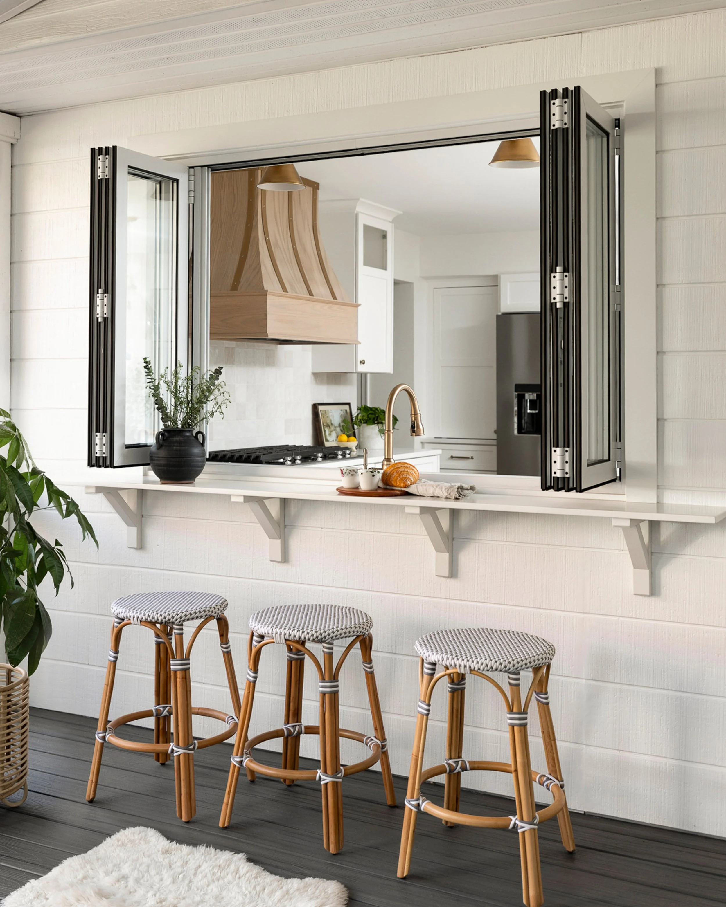 A kitchen viewed through an open window with a breakfast bar. Four wicker bar stools are lined up outside, and the kitchen has white cabinets, a wooden range hood, a stainless steel refrigerator, and a black pot with green plants on the counter.
