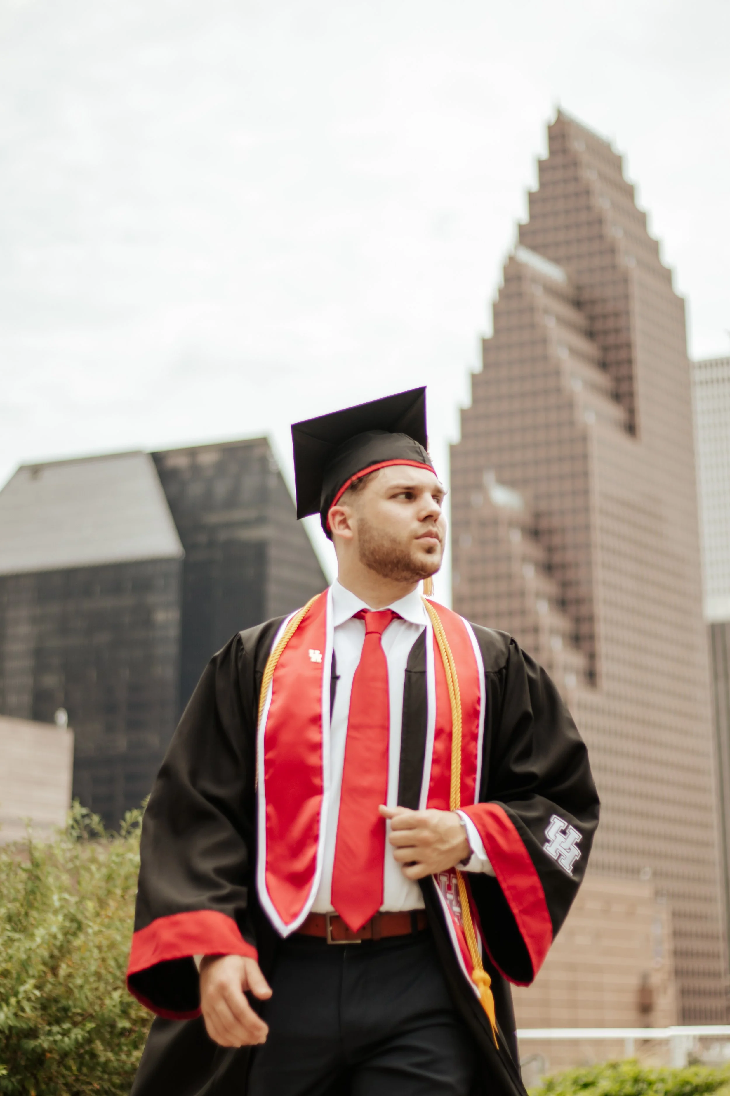 A young man in graduation attire with a black cap and gown, a red and white stole, and a red tie, standing outdoors in an urban setting with tall buildings in the background.