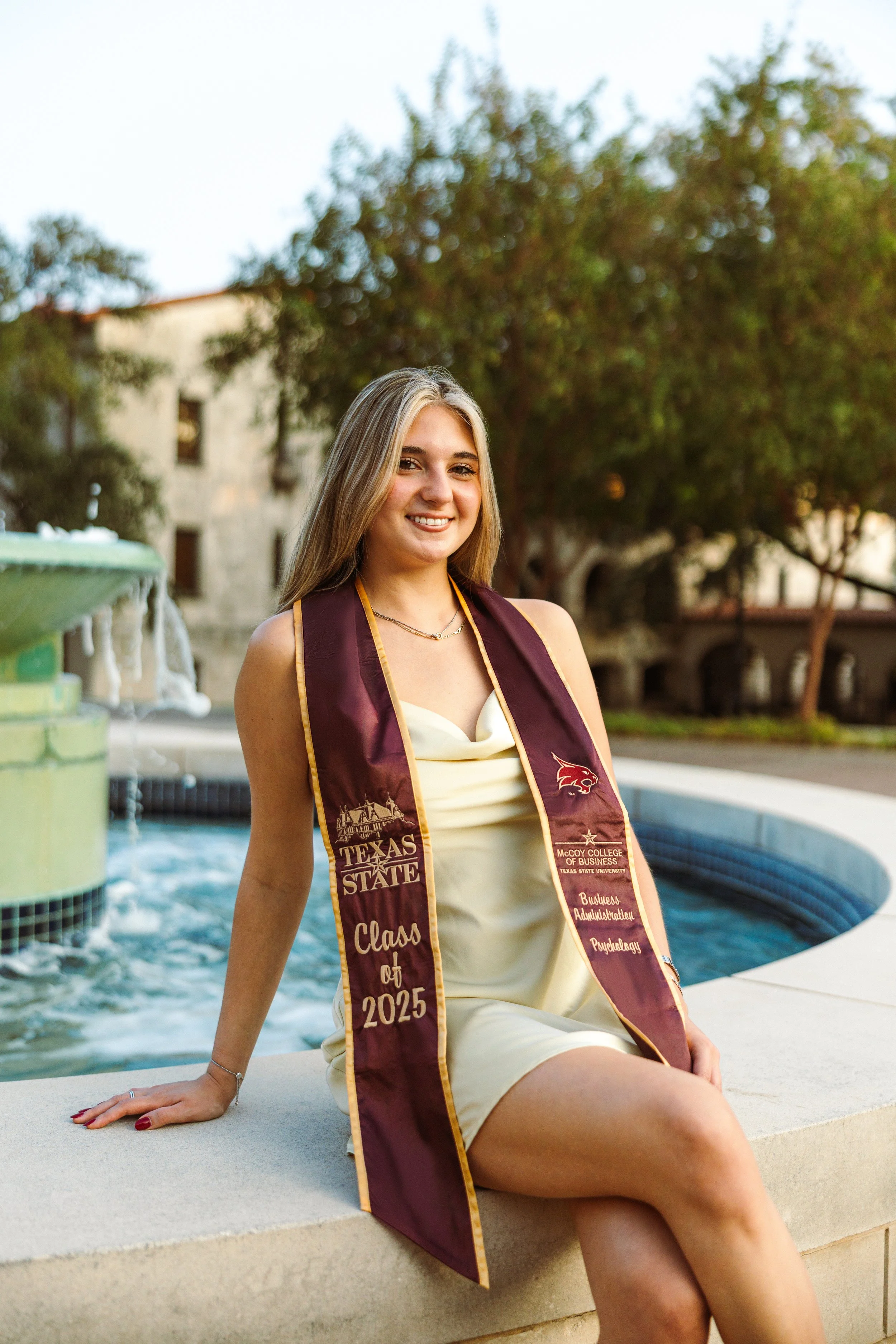 A young woman with blonde hair in a cream dress smiling and sitting on a fountain during her graduation day, wearing a maroon stole with gold trim that says 'Class of 2025' and includes logos for Texas State and McCoy College of Business.
