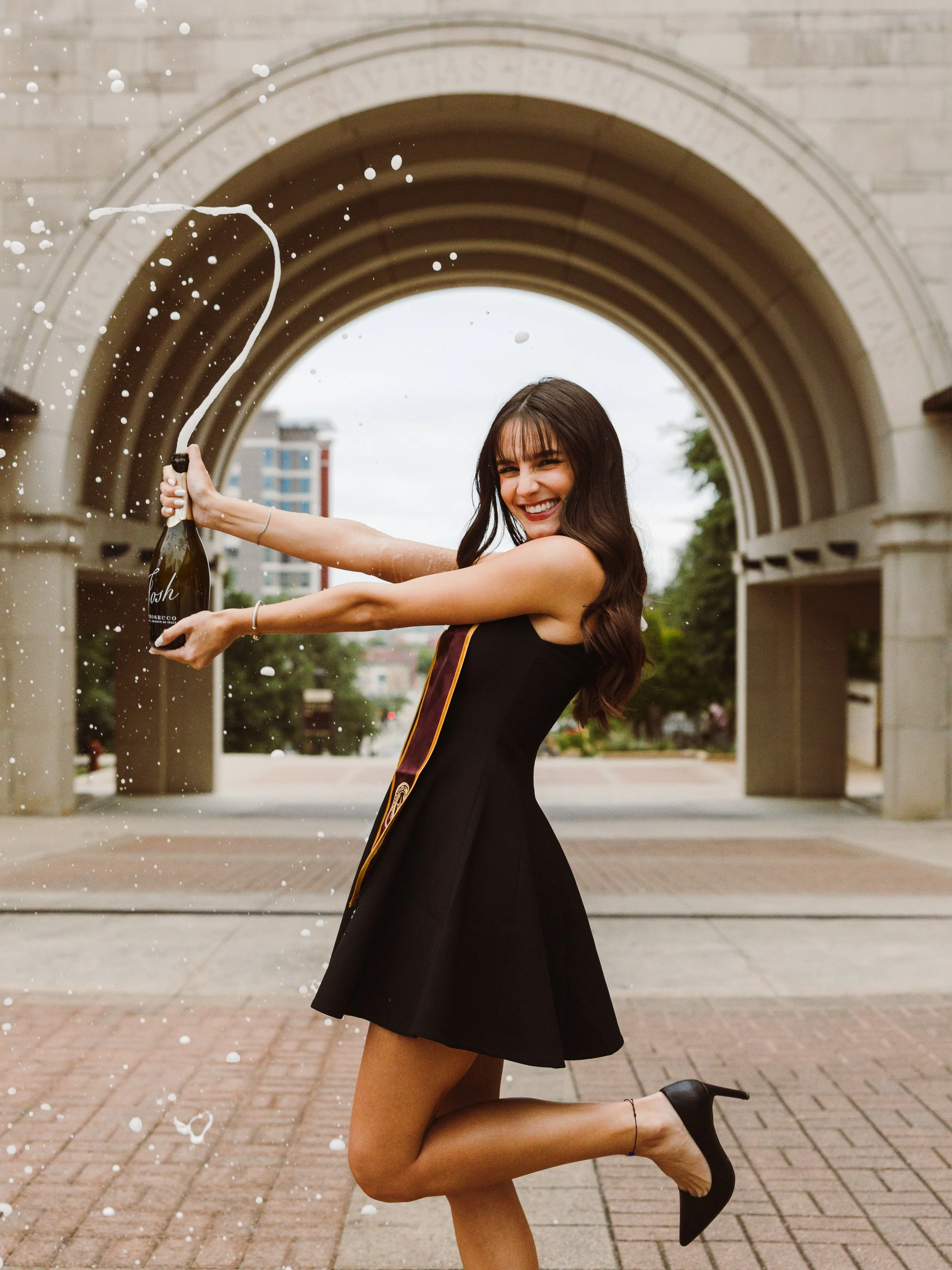 A woman in a black dress and high heels celebrates by opening a champagne bottle outdoors, with champagne spraying out, under an arched stone structure, smiling cheerfully.