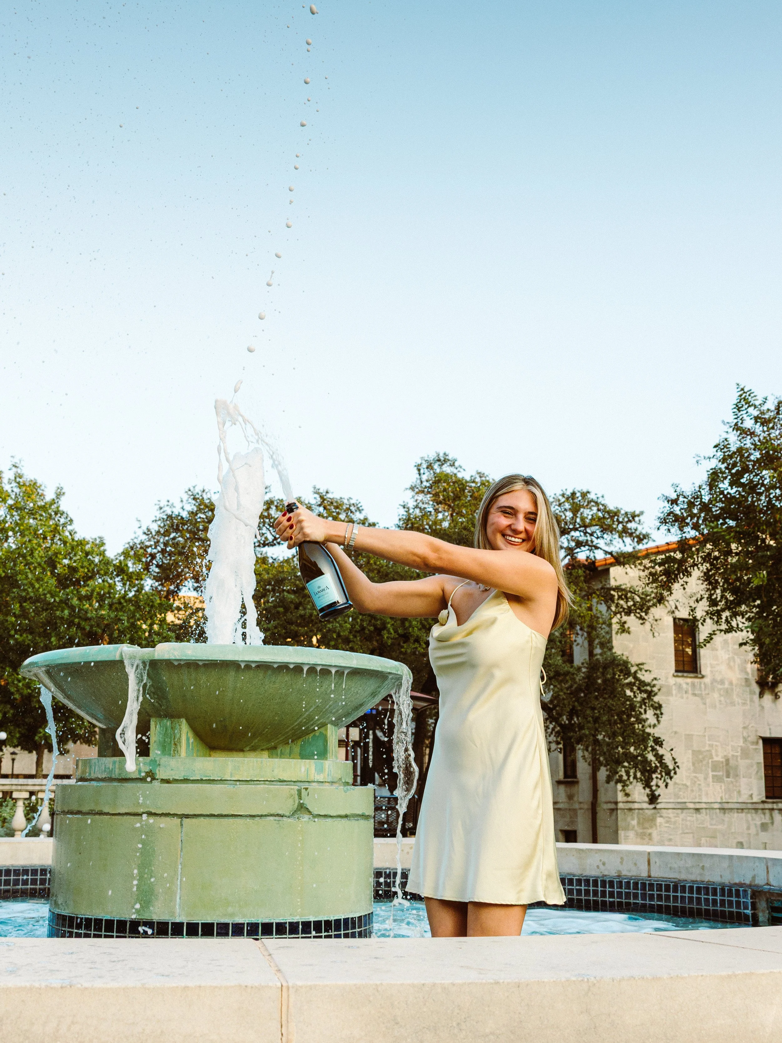 A woman in a light-colored dress celebrating with a bottle of champagne near a fountain, smiling and spraying champagne in the air, outdoors with trees and a building in the background.