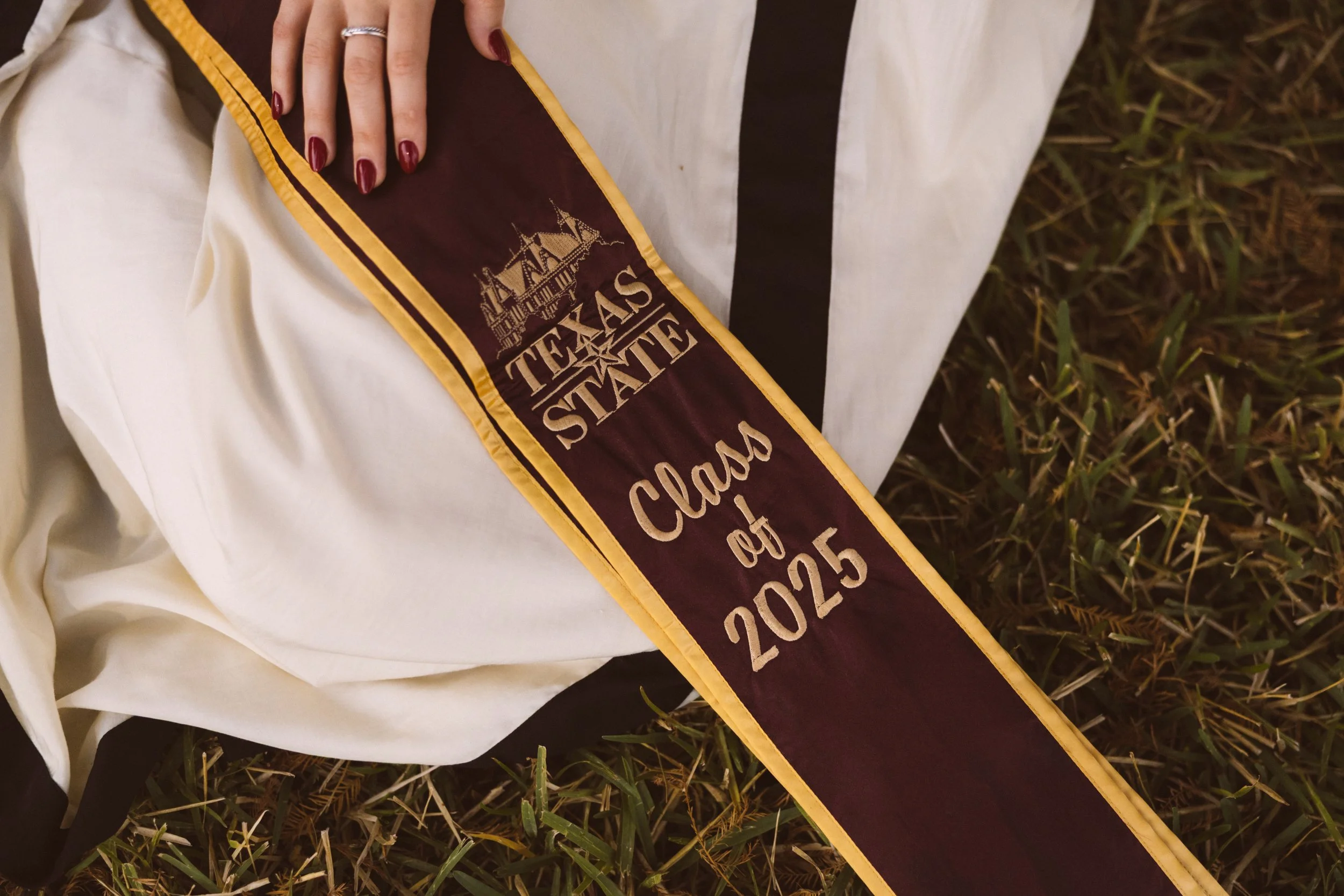A graduation sash with gold trim, reading 'Texas State Class of 2025', placed on grass.