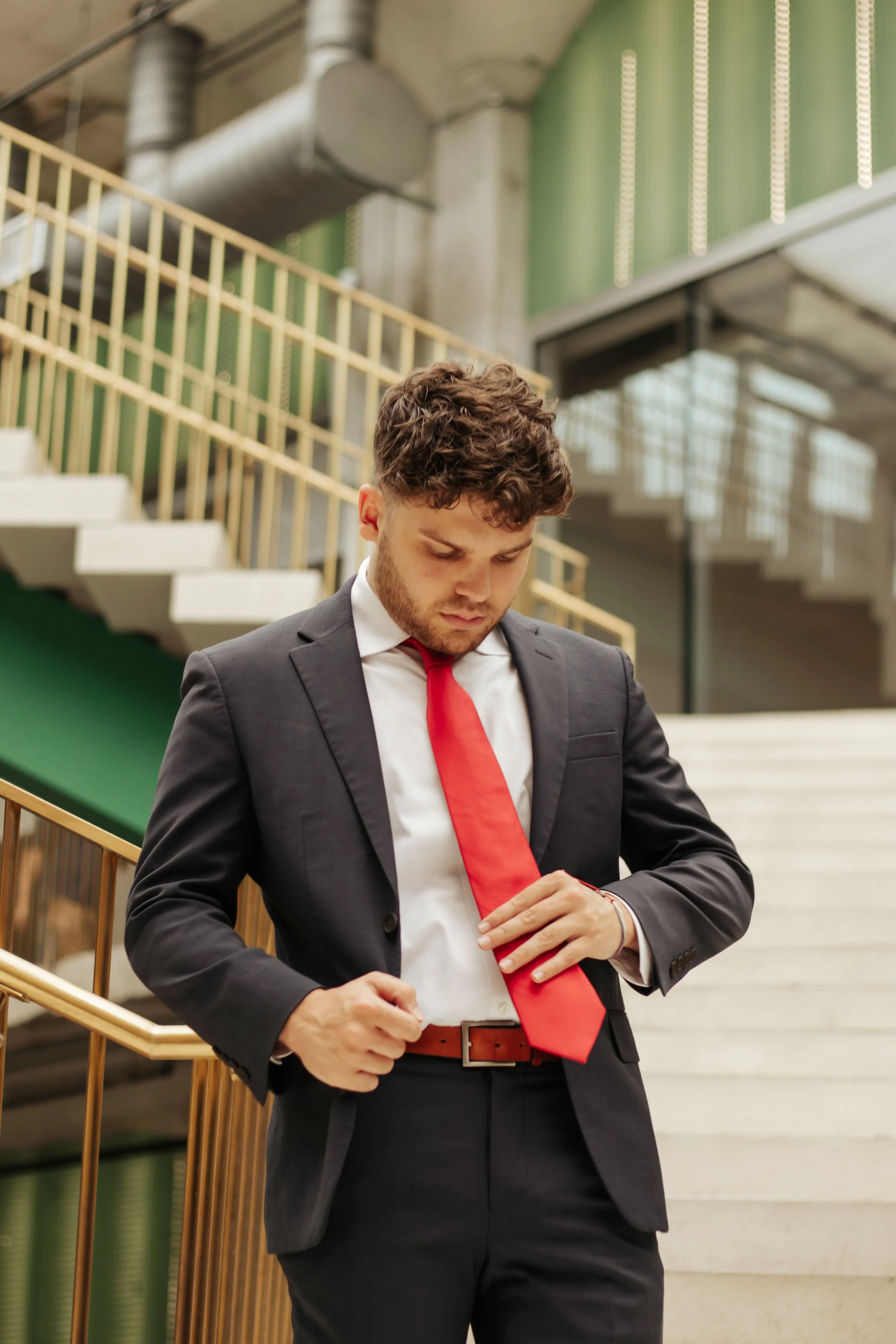 A young man in a black suit with a white shirt and red tie stands on a staircase in an industrial-style building, adjusting his belt or jacket.