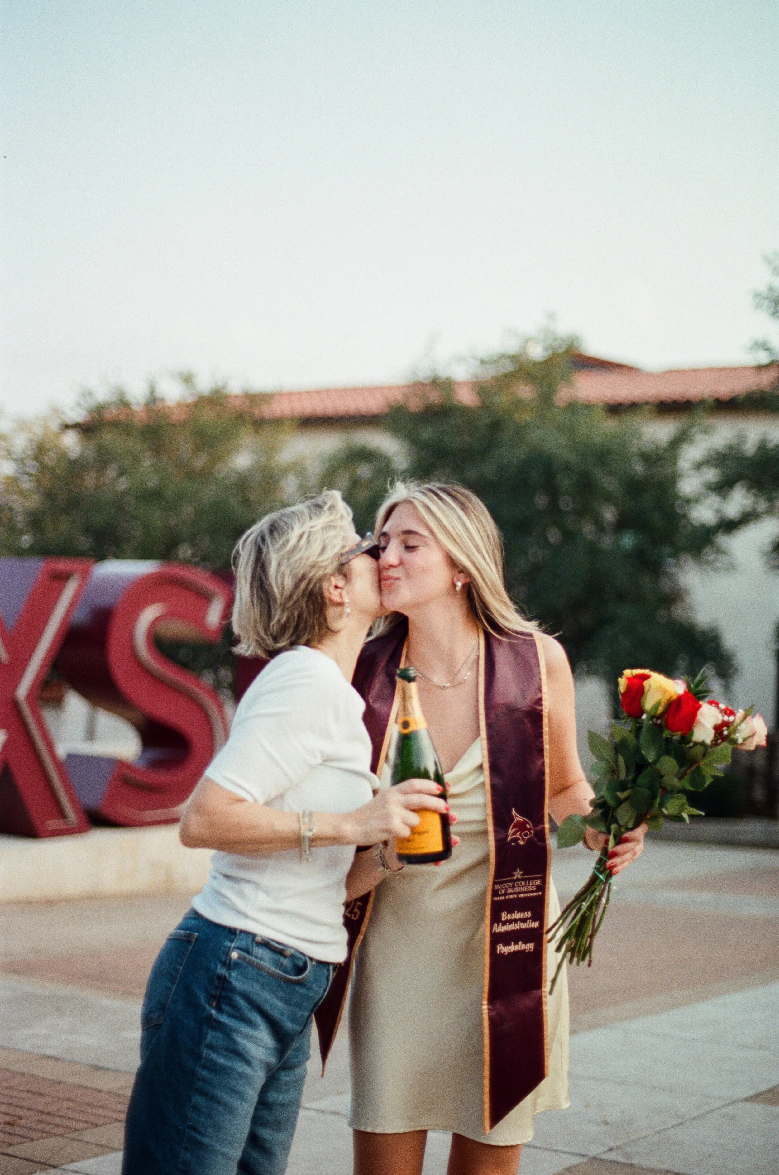 A woman with a graduation stole kisses an older woman who is holding a bouquet of roses and a champagne bottle, celebrating graduation outdoors.