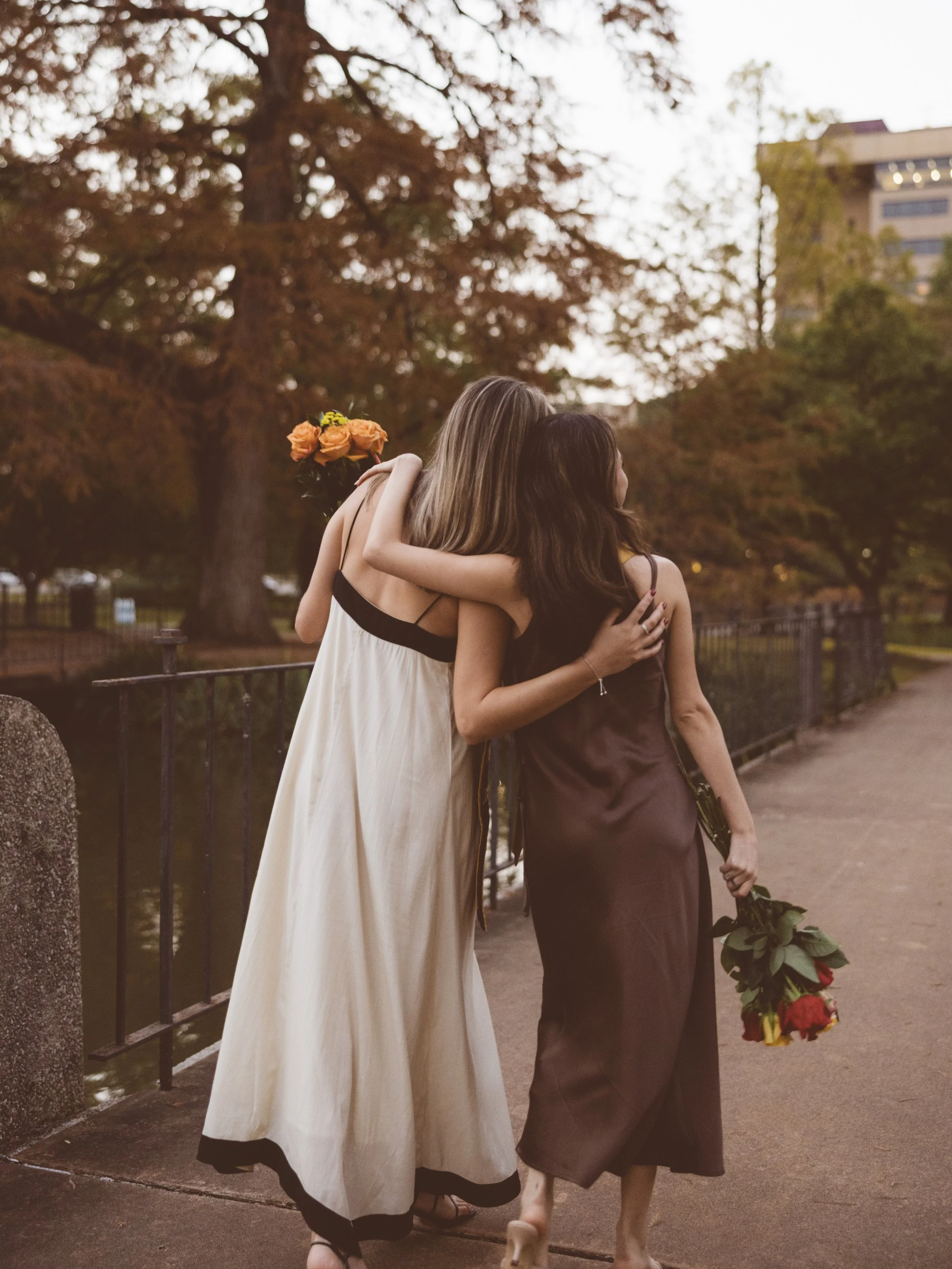 Two women hug each other, one holding a bouquet of roses, during sunset in a park.