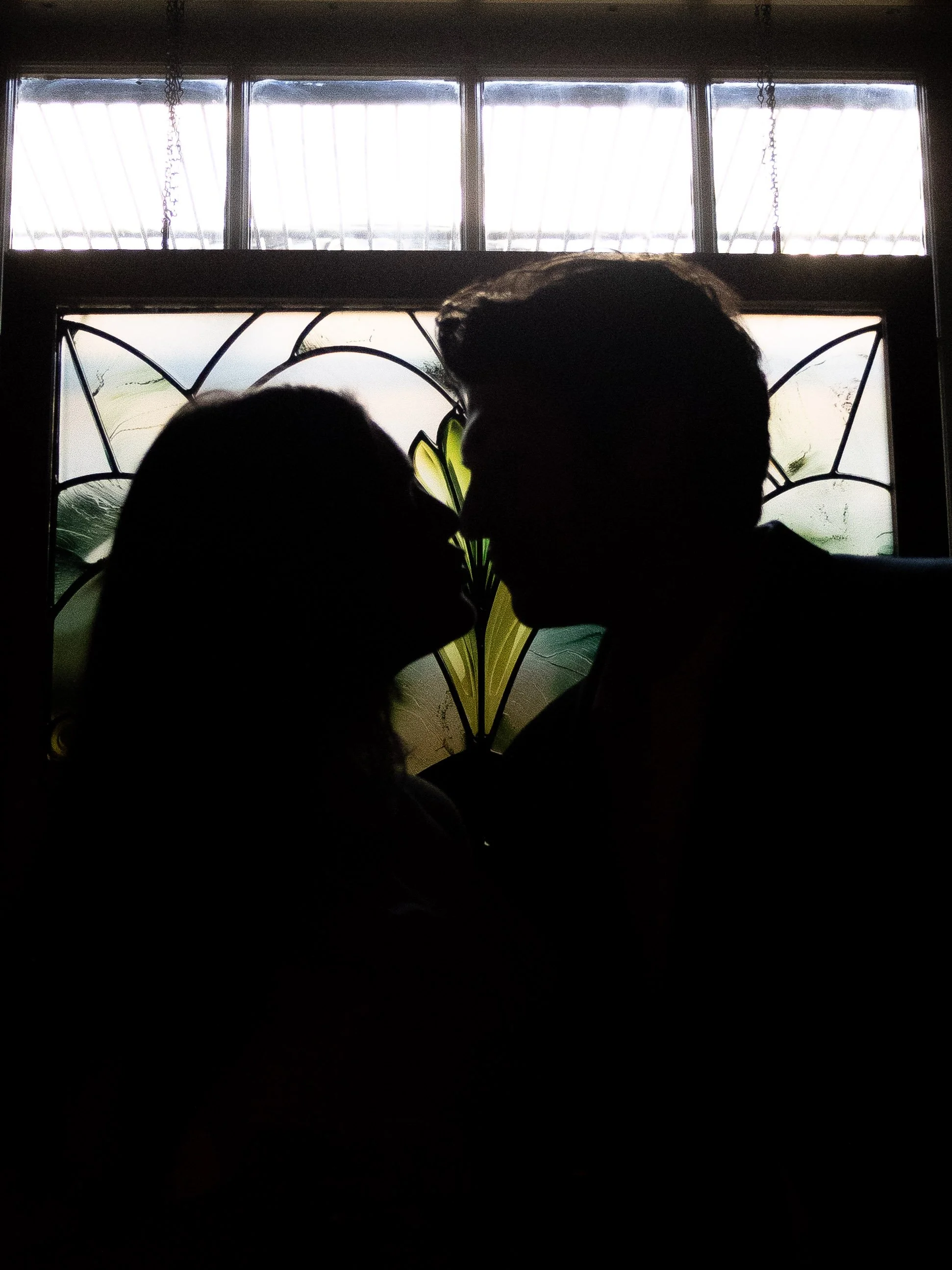Silhouettes of a couple about to kiss in front of a decorative window with stained glass design.