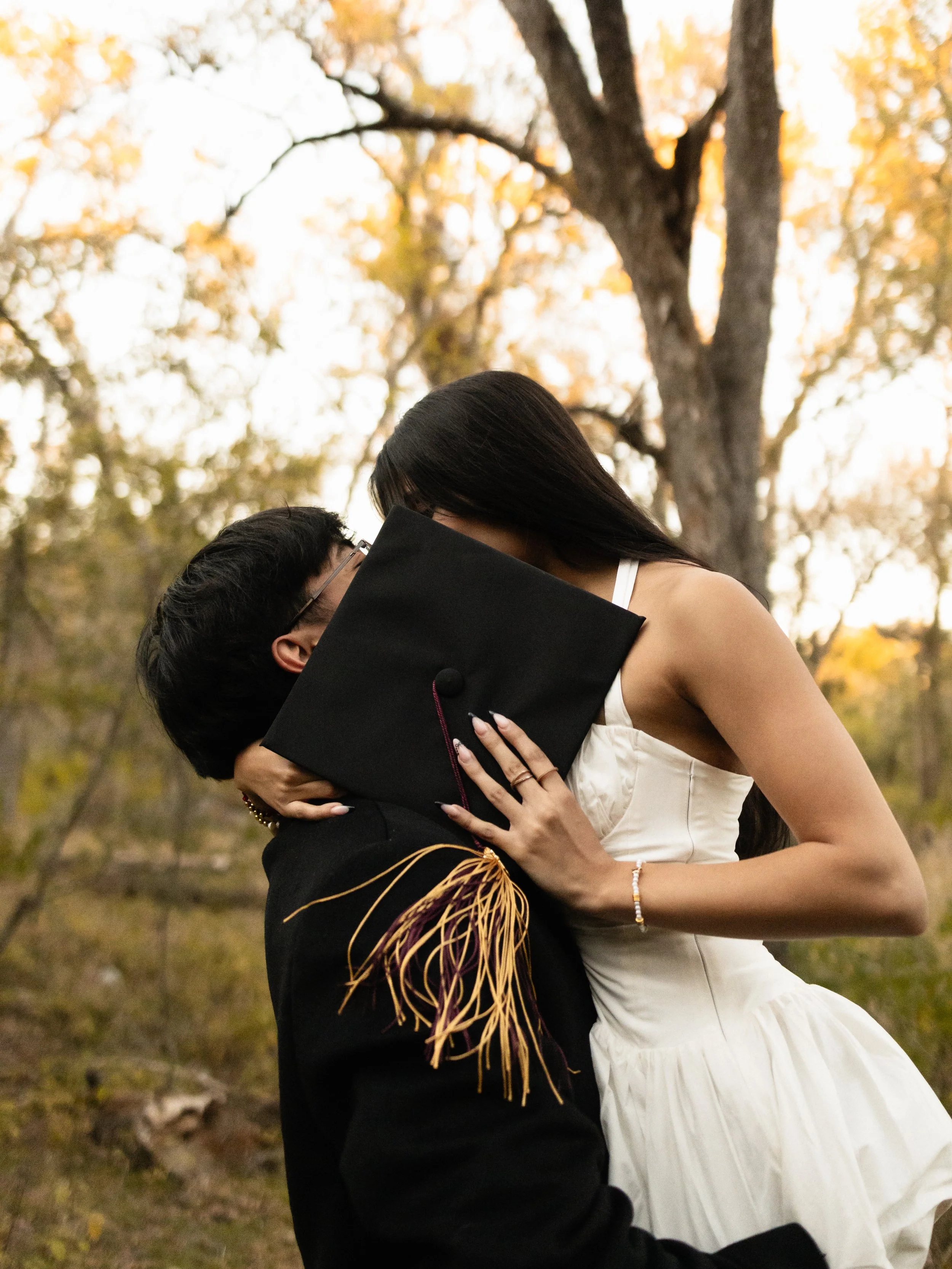 A couple in a kiss outdoors during autumn, with the person in a cap and gown holding a graduation cap, and the woman in a white dress.