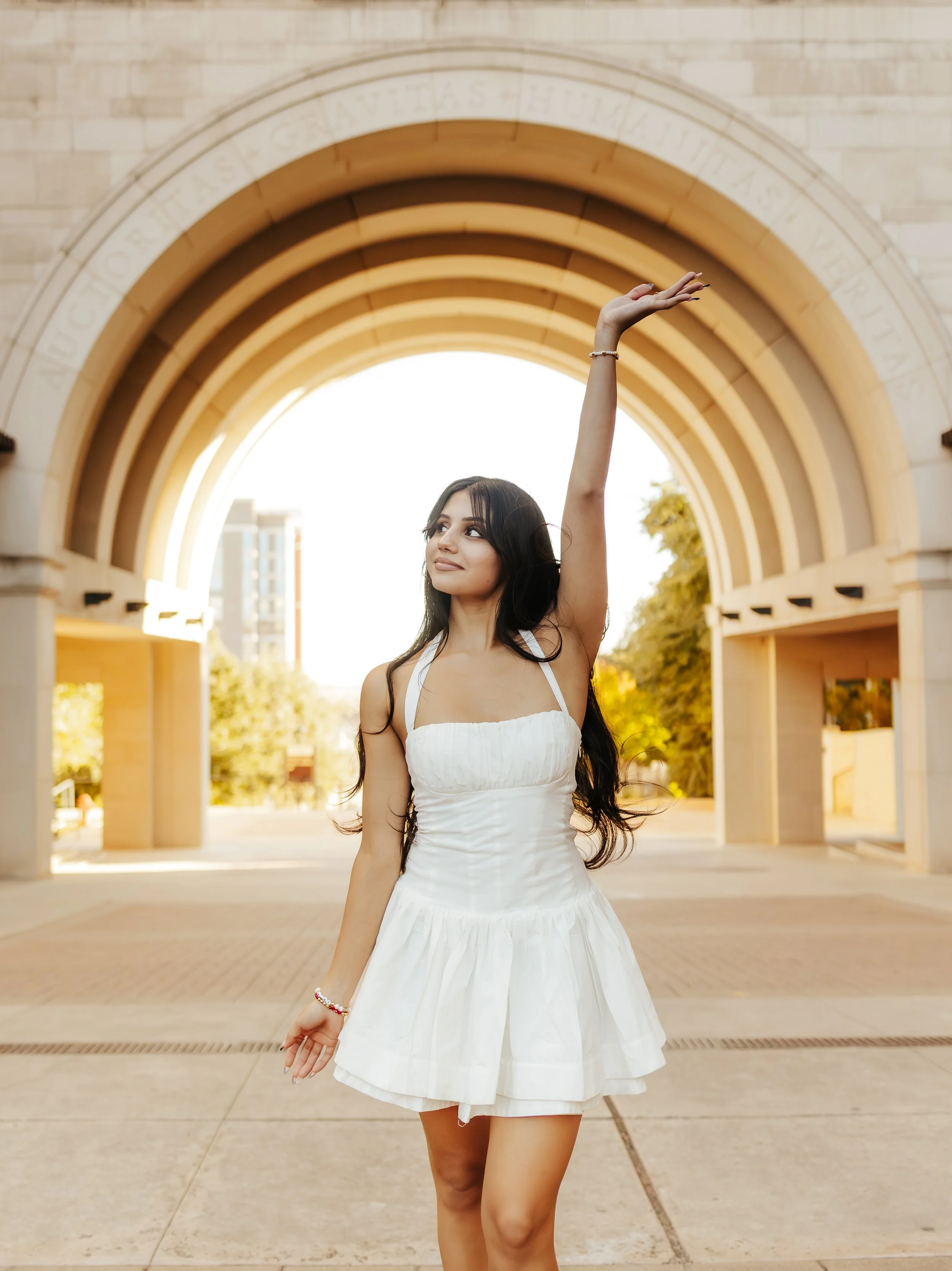 A woman in a white dress standing outdoors under an archway with her right arm raised in a graceful pose.