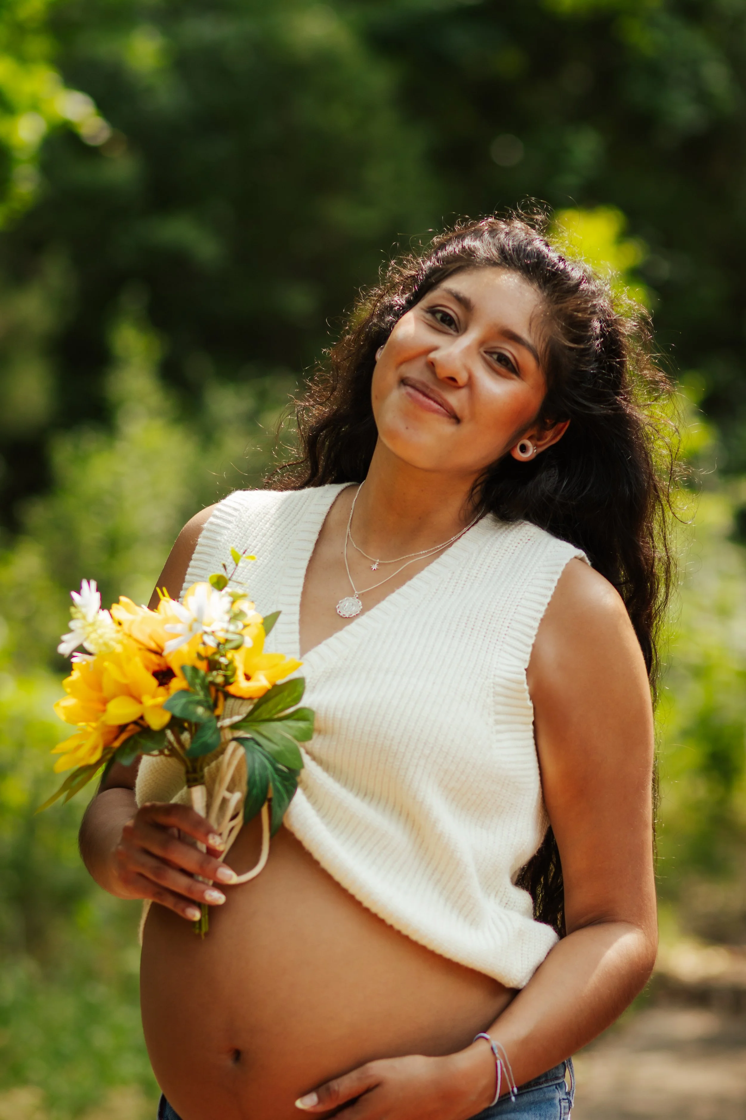 A pregnant woman with long, wavy black hair outdoors, holding a bouquet of yellow and white flowers, smiling and wearing a white sleeveless sweater and layered necklaces.