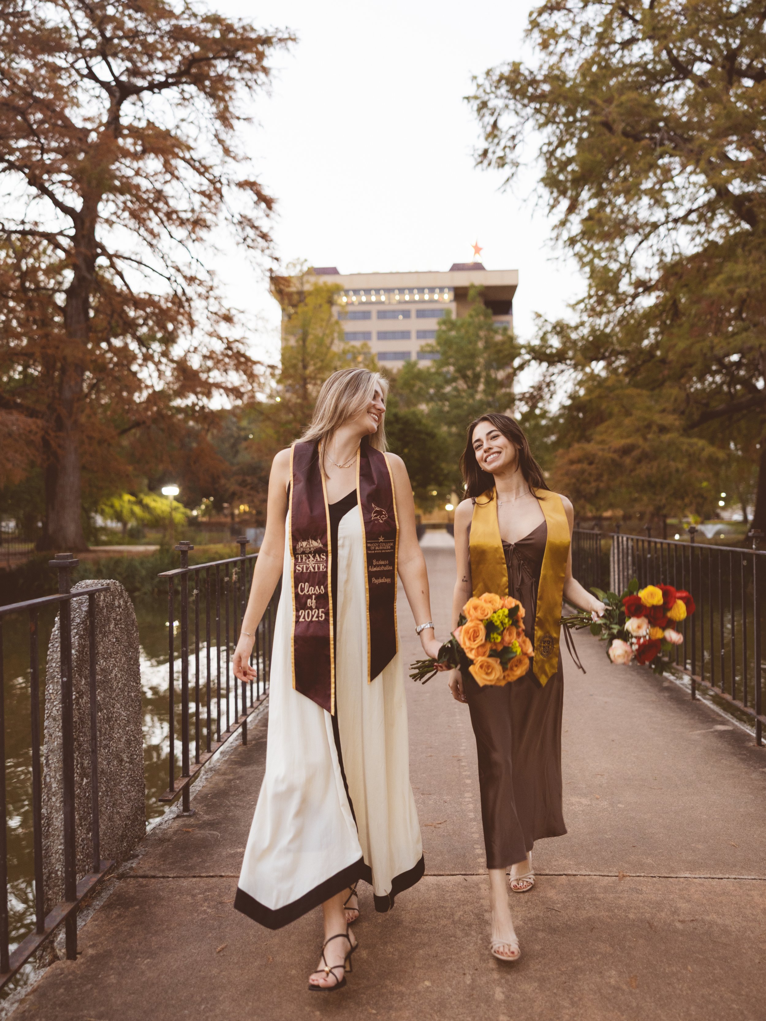Two women wearing graduation stoles walk hand-in-hand on a bridge while holding bouquets of flowers, smiling, with trees and a building in the background.