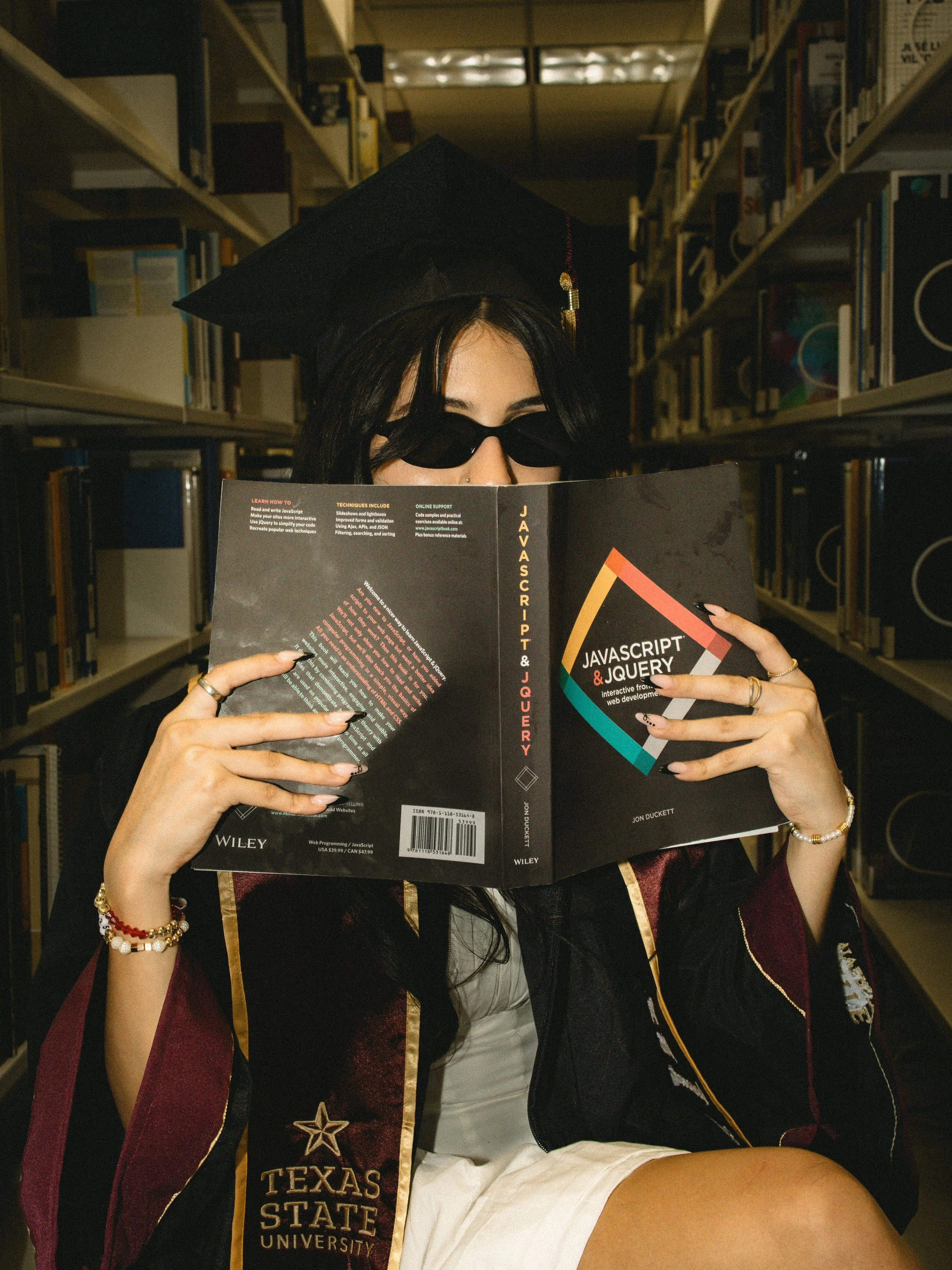 A graduate wearing a cap, gown, and sunglasses sitting in a library, reading a book titled 'JavaScript & JQuery'. The graduate has long dark hair, multiple rings and bracelets, and is holding the book with both hands. The library shelves behind are f
