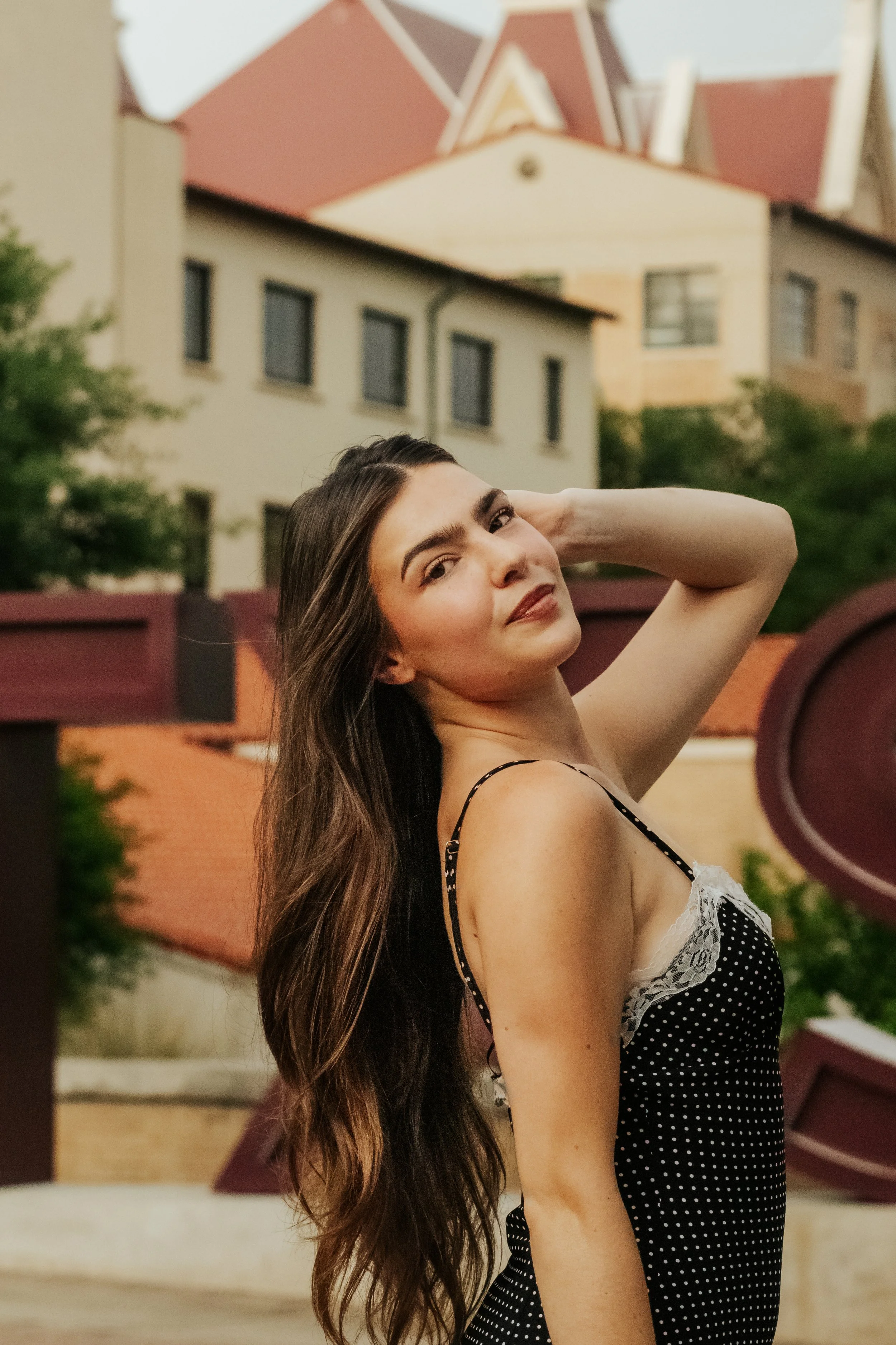 A young woman with long brown hair, wearing a black polka dot dress with lace details, stands outdoors with her hand resting behind her head, looking at the camera against a backdrop of buildings and trees.