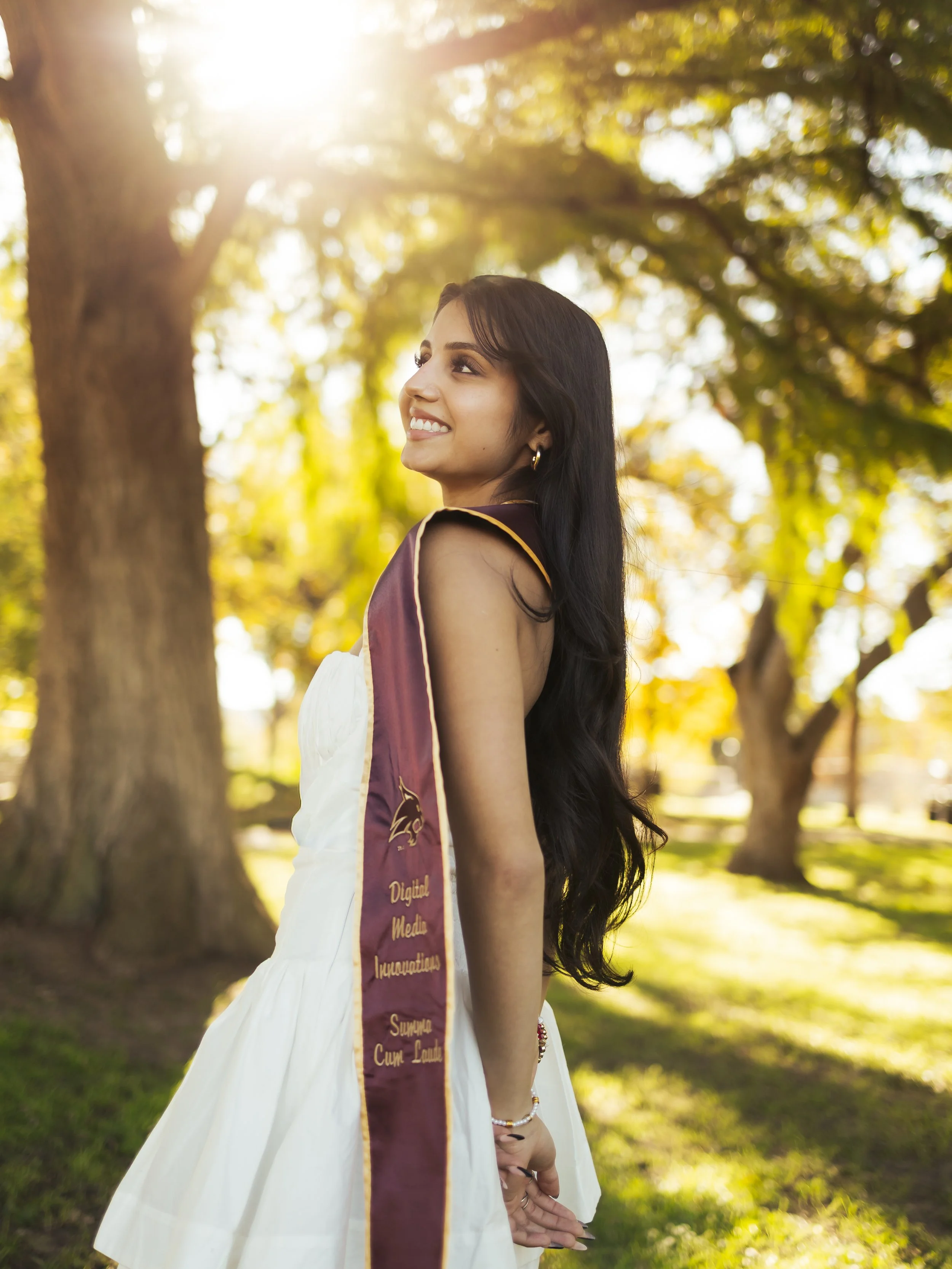 Young woman in white dress with maroon sash standing outdoors in a park with trees, sunlight, and green grass, smiling and looking to her left.