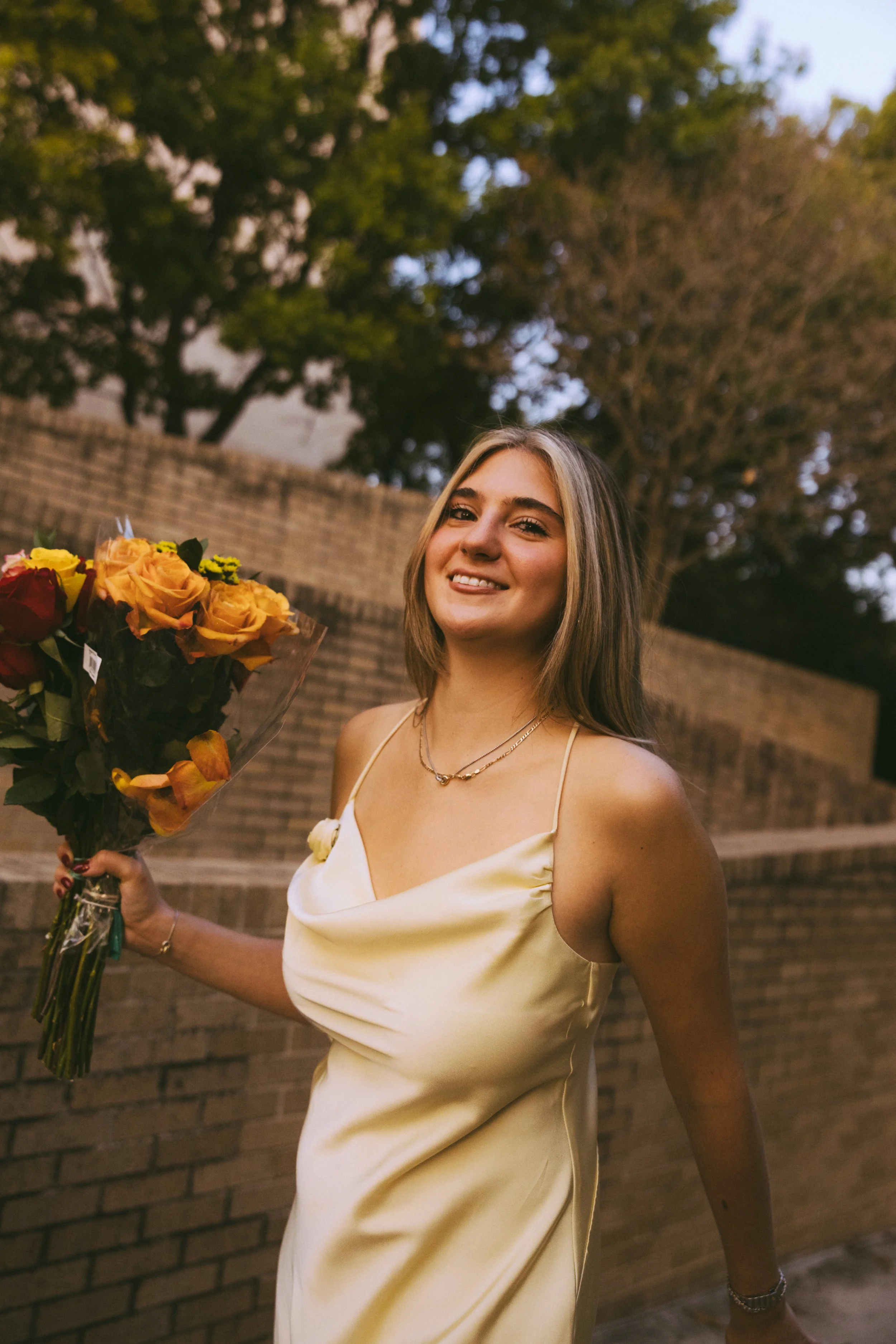A young woman with shoulder-length blonde hair wearing a silky yellow dress, holding a bouquet of colorful flowers, smiling outdoors with trees and a brick wall in the background.