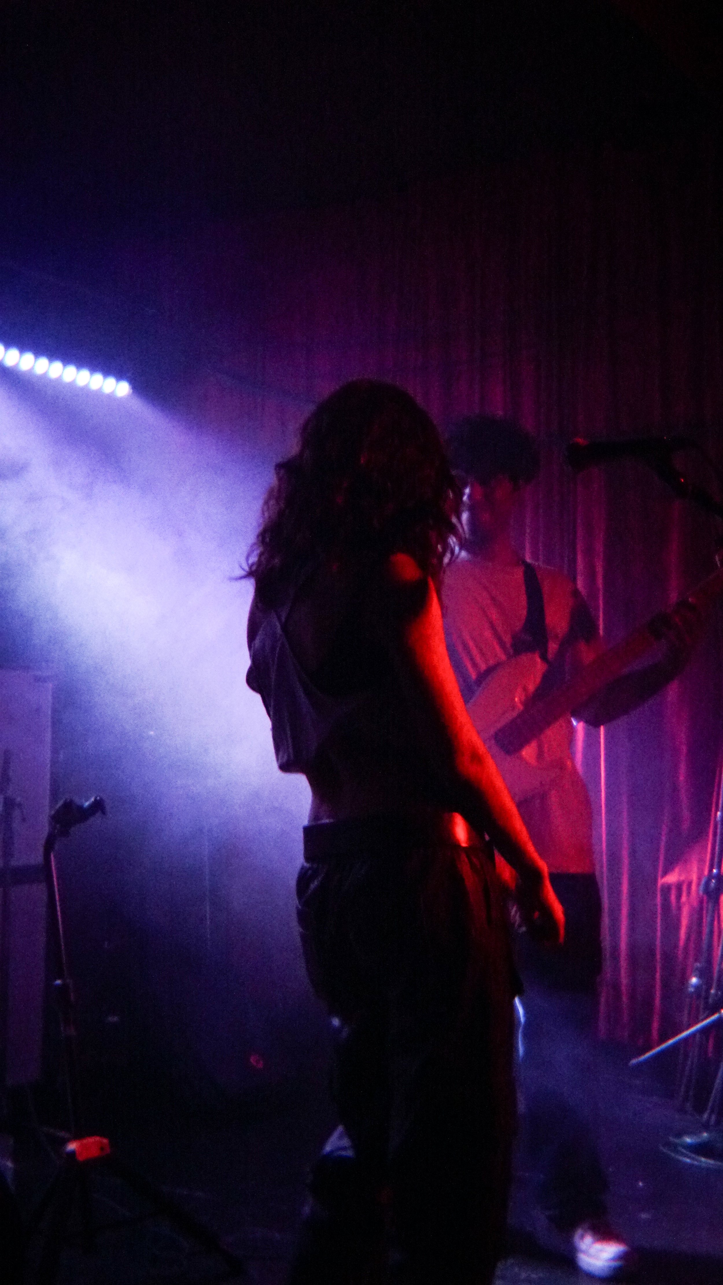 A woman with curly hair stands on stage in front of a microphone, while a man plays guitar behind her. The scene is dark with colorful stage lighting creating a vibrant, atmospheric setting.