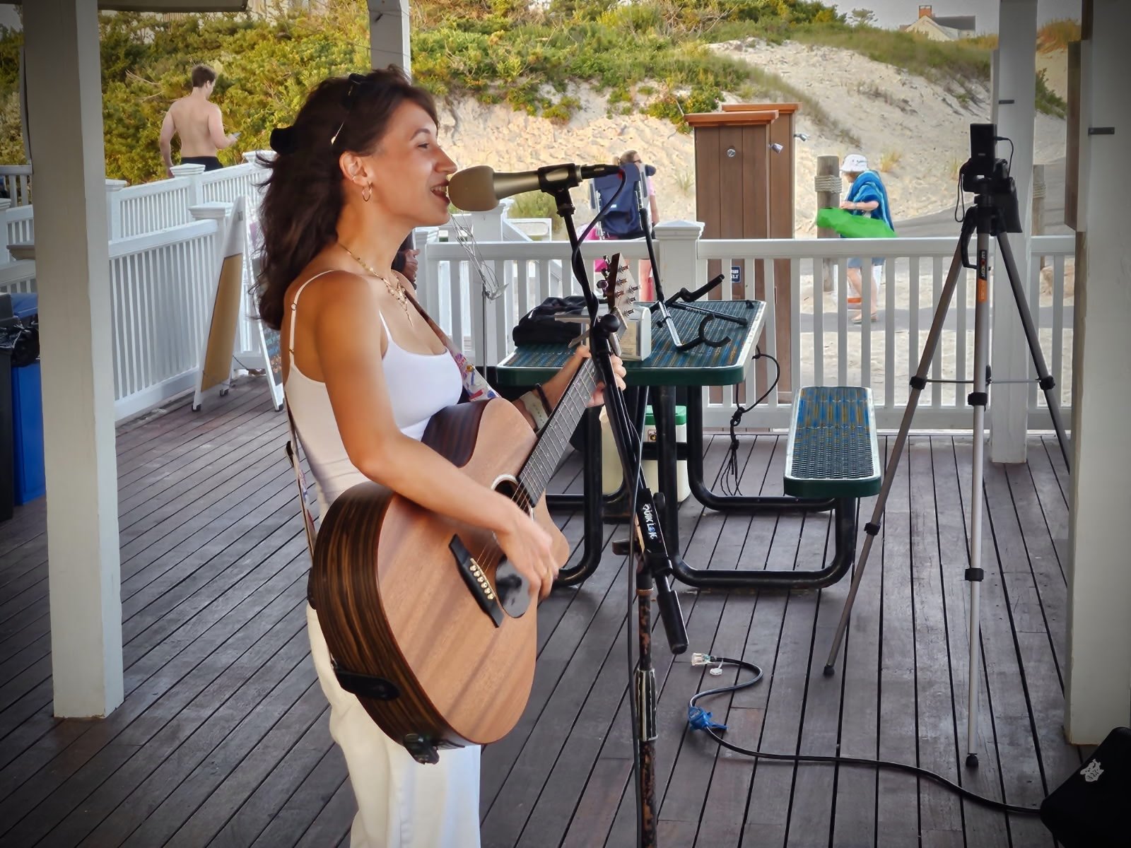 A woman singing into a microphone and playing an acoustic guitar at an outdoor venue with a coastal background. Two tripods with equipment and a bench are visible in the scene.