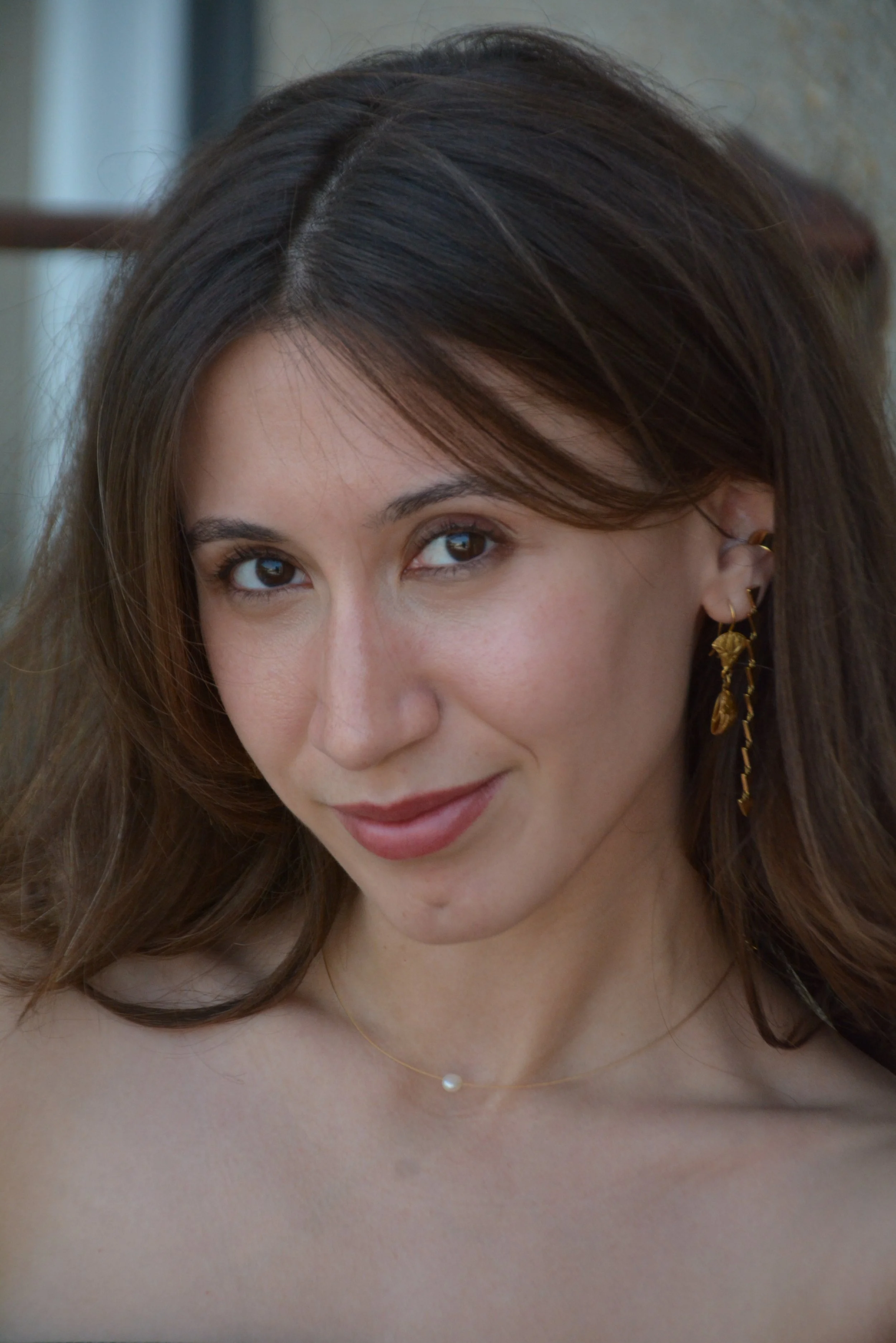 Close-up of a woman with brown hair and brown eyes, smiling slightly, wearing earrings and a delicate necklace.