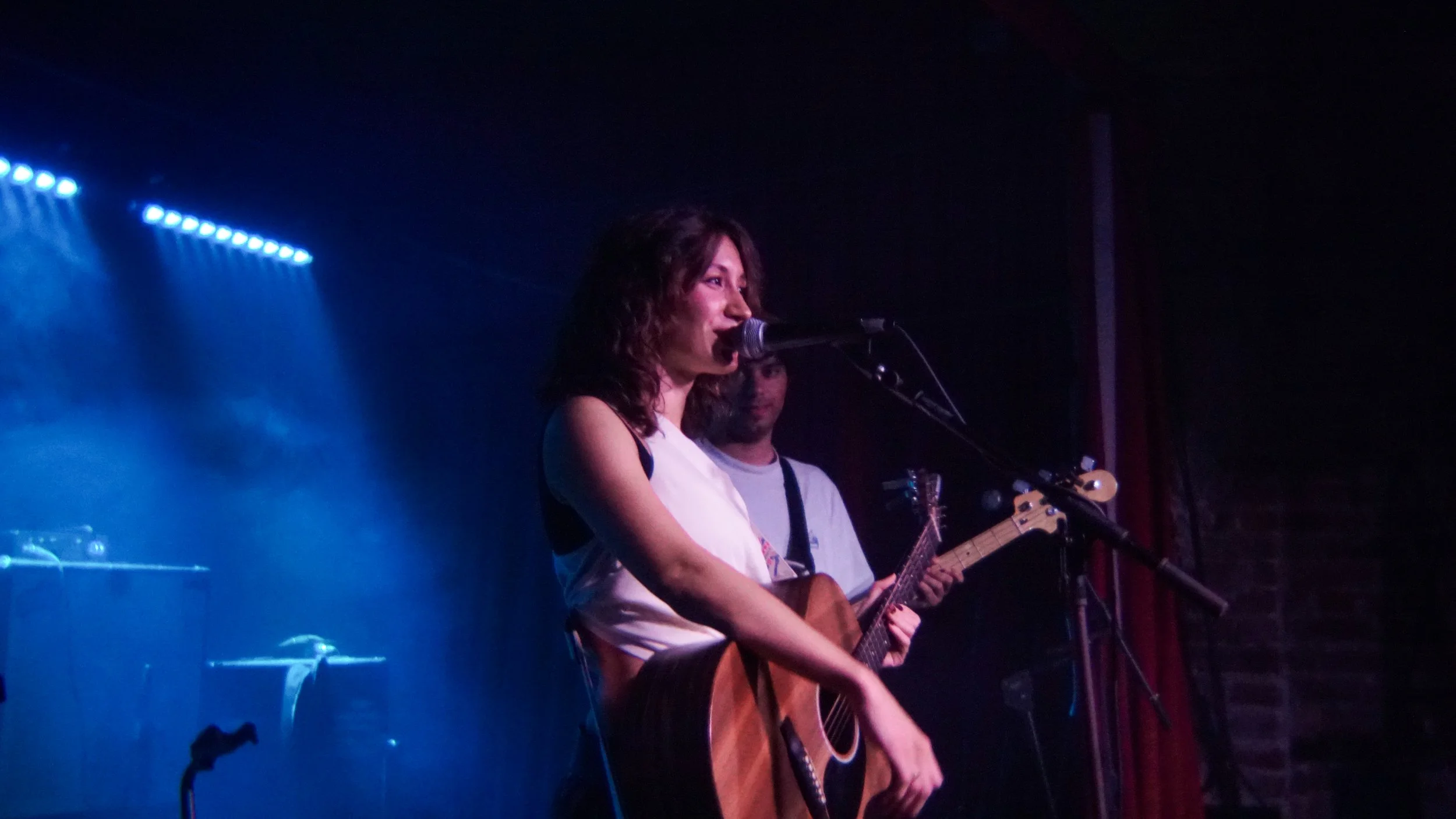 A woman with brown hair singing into a microphone while playing an acoustic guitar on stage, with a man in the background playing an electric bass.