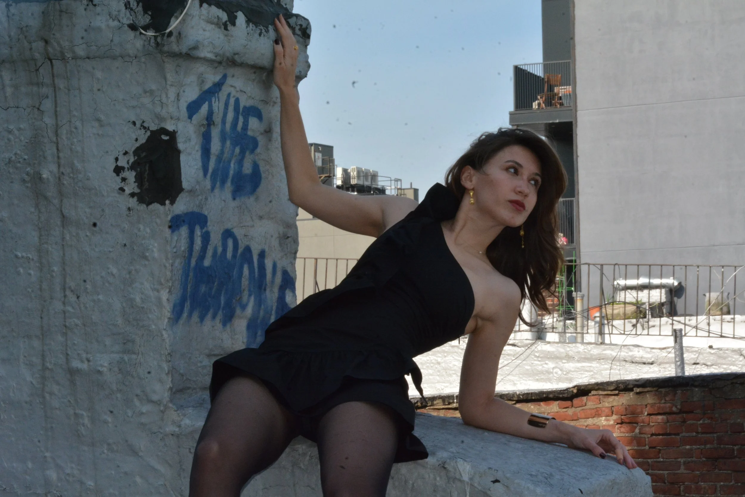 A woman in a black dress and sheer stockings reclines on a rooftop ledge, with urban buildings and incomplete construction visible in the background.