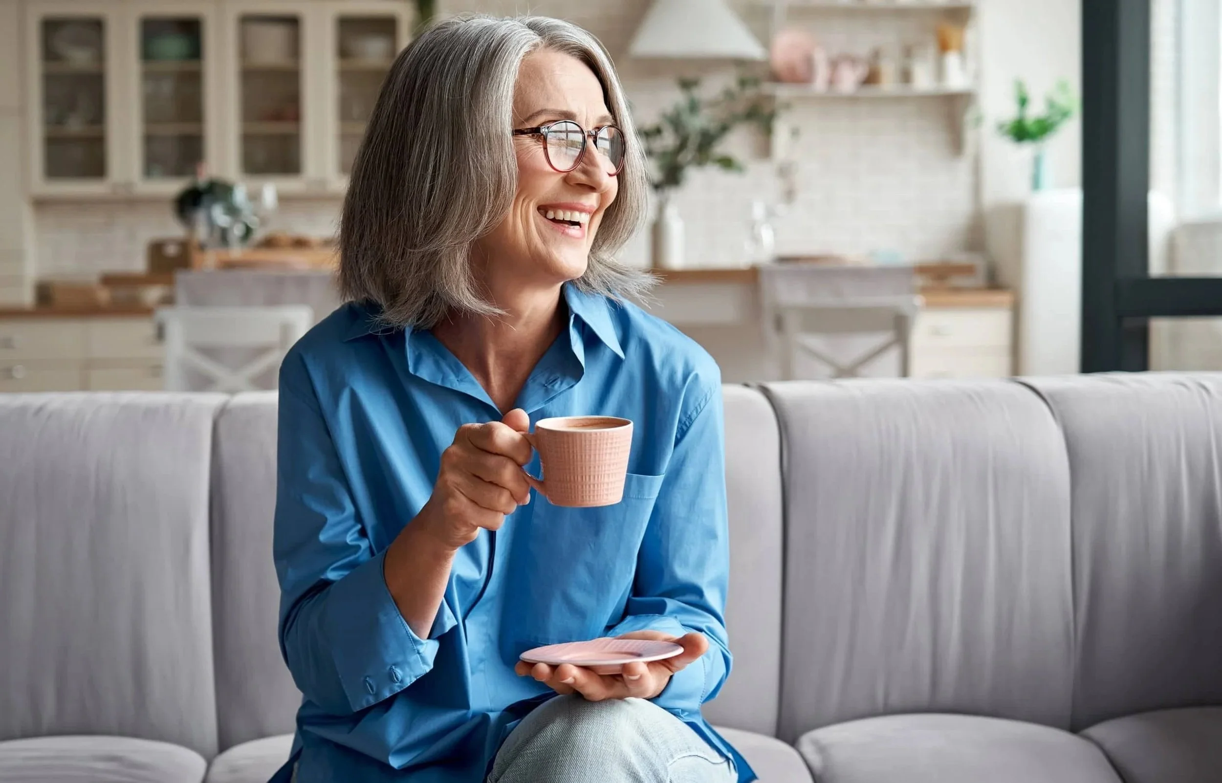 A mature woman with gray hair and glasses, wearing a blue shirt, sitting on a light-colored sofa in a bright kitchen, holding a pink cup and smiling.