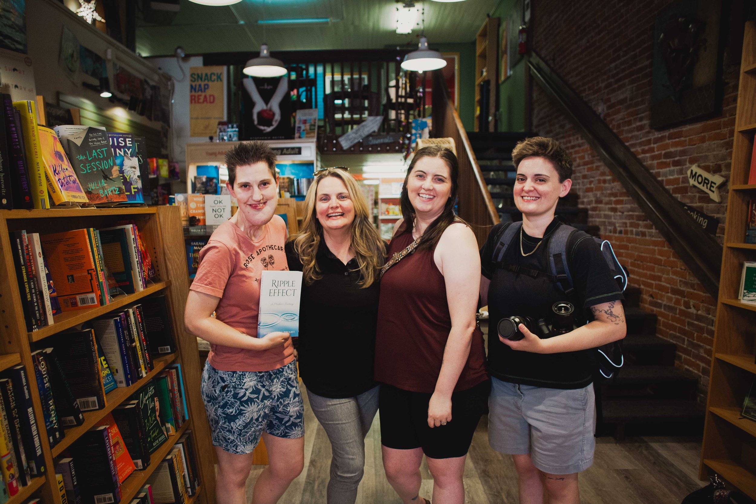 Group portrait of attendees and photographers smiling together inside Pearl Street Books during a Ripple Effect author event and book signing.