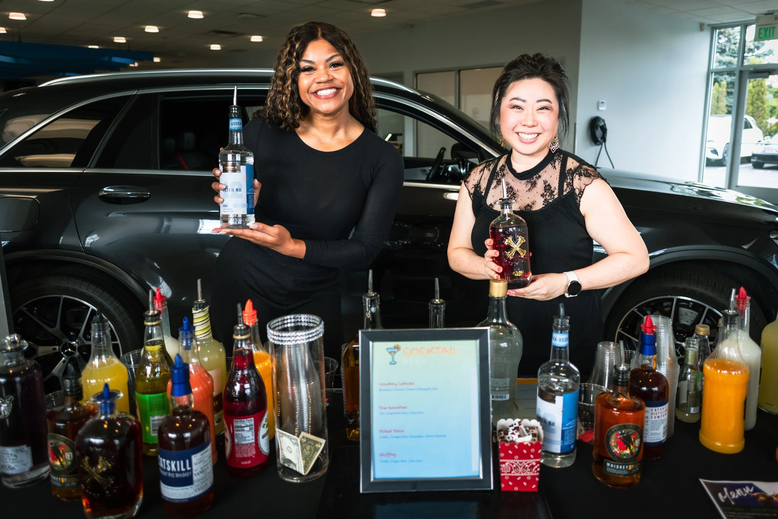 two smiling women sit behind a mobile cocktail bar display inside a car show room. Each woman holds a bottle of liquor while standing in front of a black vehicle . the table infron tof them is filled with cocktail ingredients,