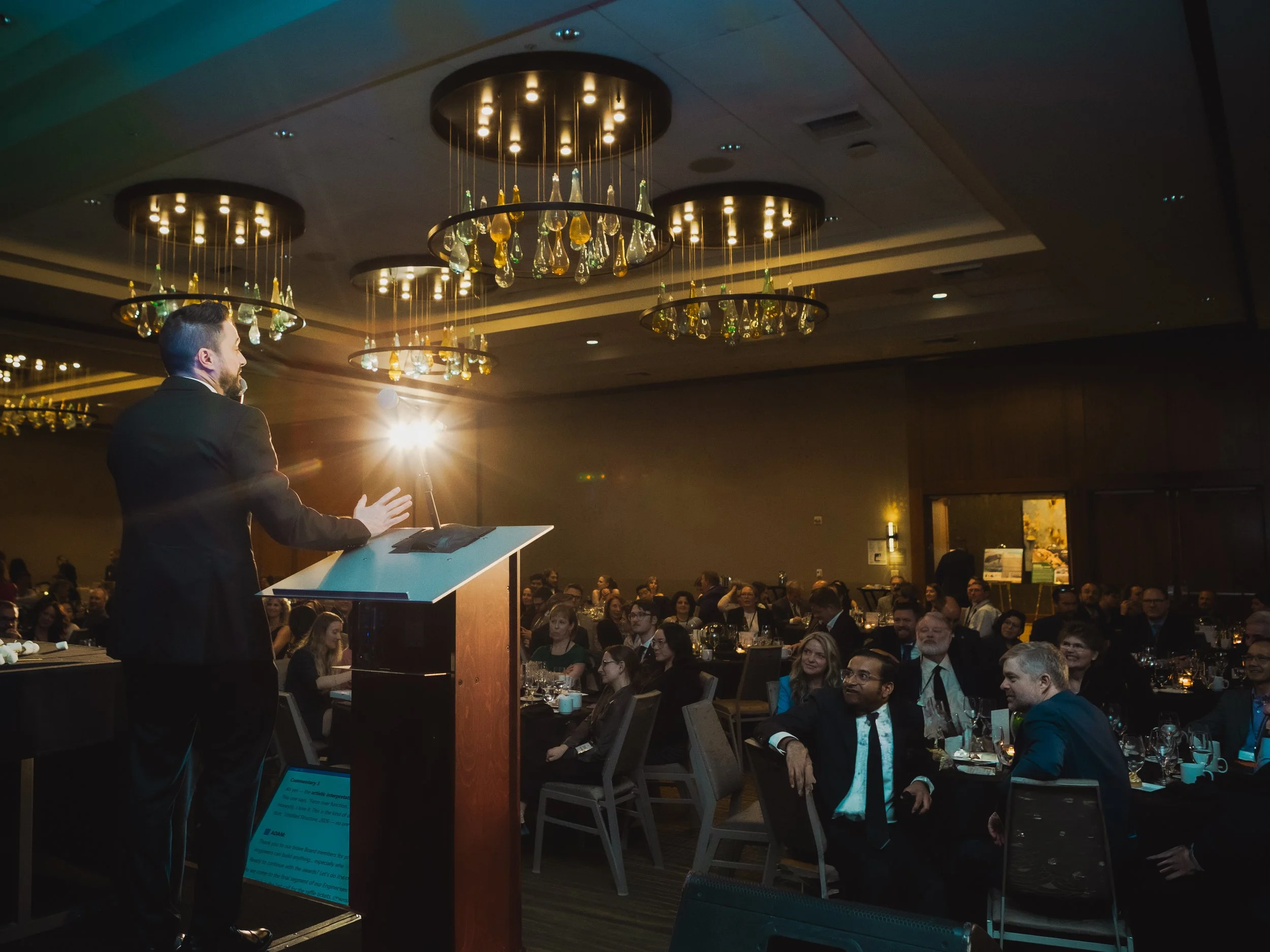 A speaker stands at a podium addressing a large audience during a formal awards or conference event. Chandeliers hang overhead while attendees listen from round banquet tables.
