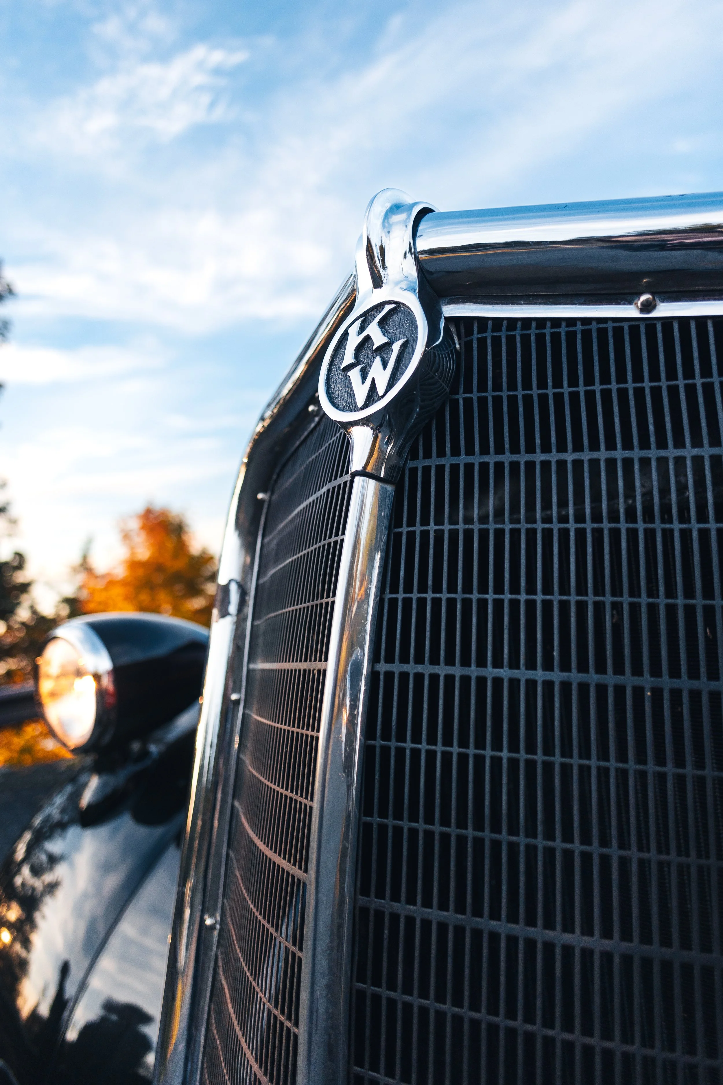 Close-up of the chrome front grille and Kenworth emblem on a vintage national park tour bus on Orcas Island, Washington, with a blue sky and autumn-colored trees softly blurred in the background.