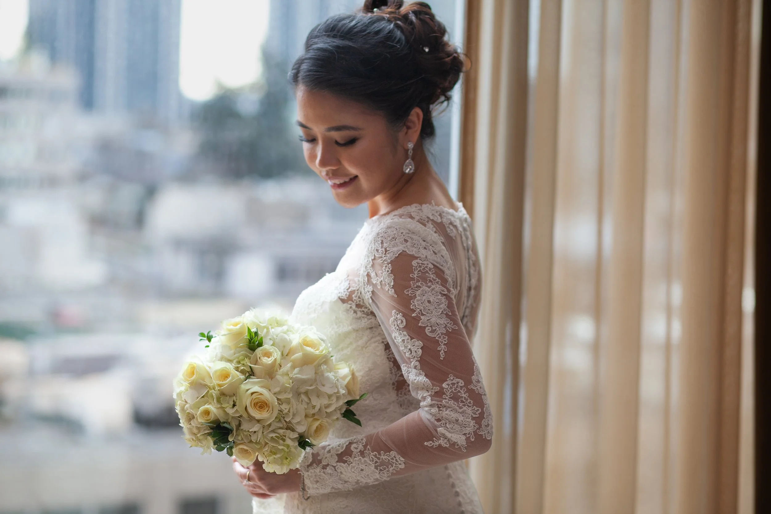 bride admires floral bouquet while standing in the window at the bellevue hyatt hotel, white roses, asian bride, lace wedding dress