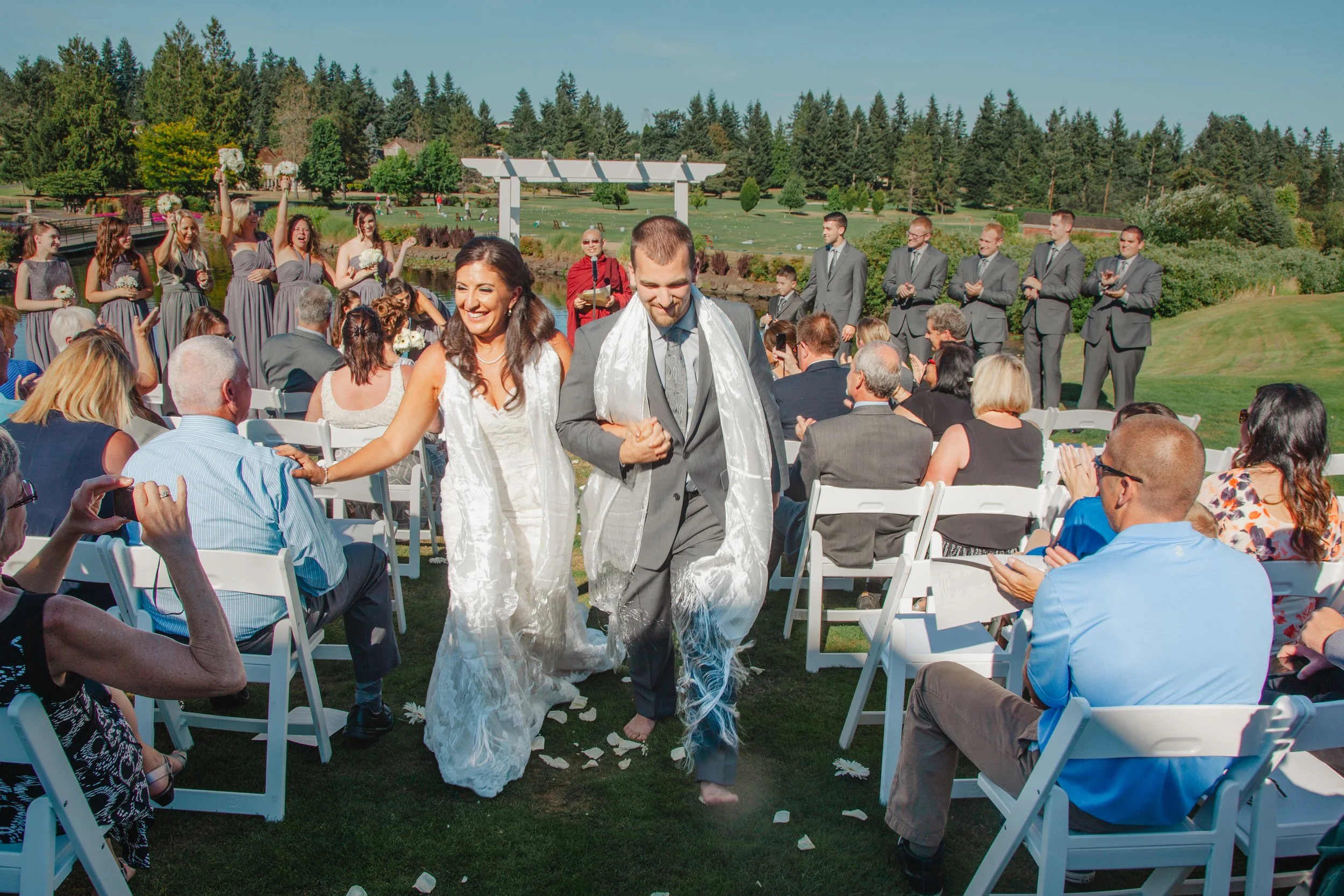 Bride and groom walking down the aisle after wedding ceremony outdoors, surrounded by seated guests, with bridal party and lawn in the background.