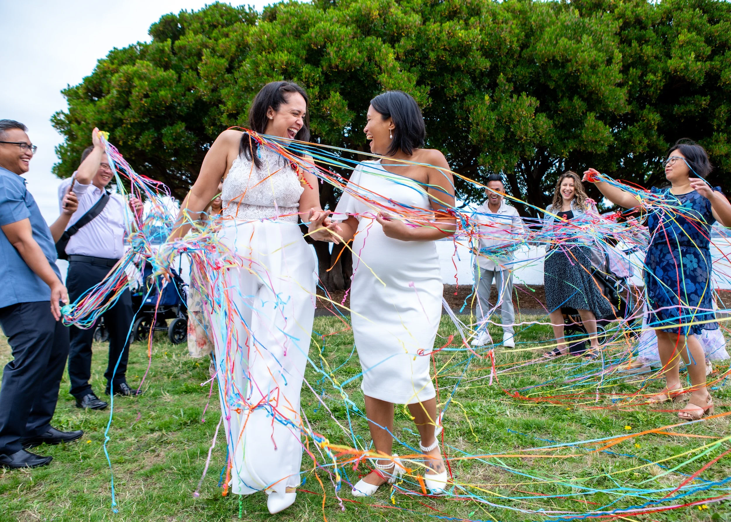 Leah and Babylynn laugh together during their wedding celebration as colorful ribbons intertwine around the couple and their guests, capturing a joyful, candid moment of their outdoor wedding. Photographed by Tayler Berard Creative in the Pacific Nor