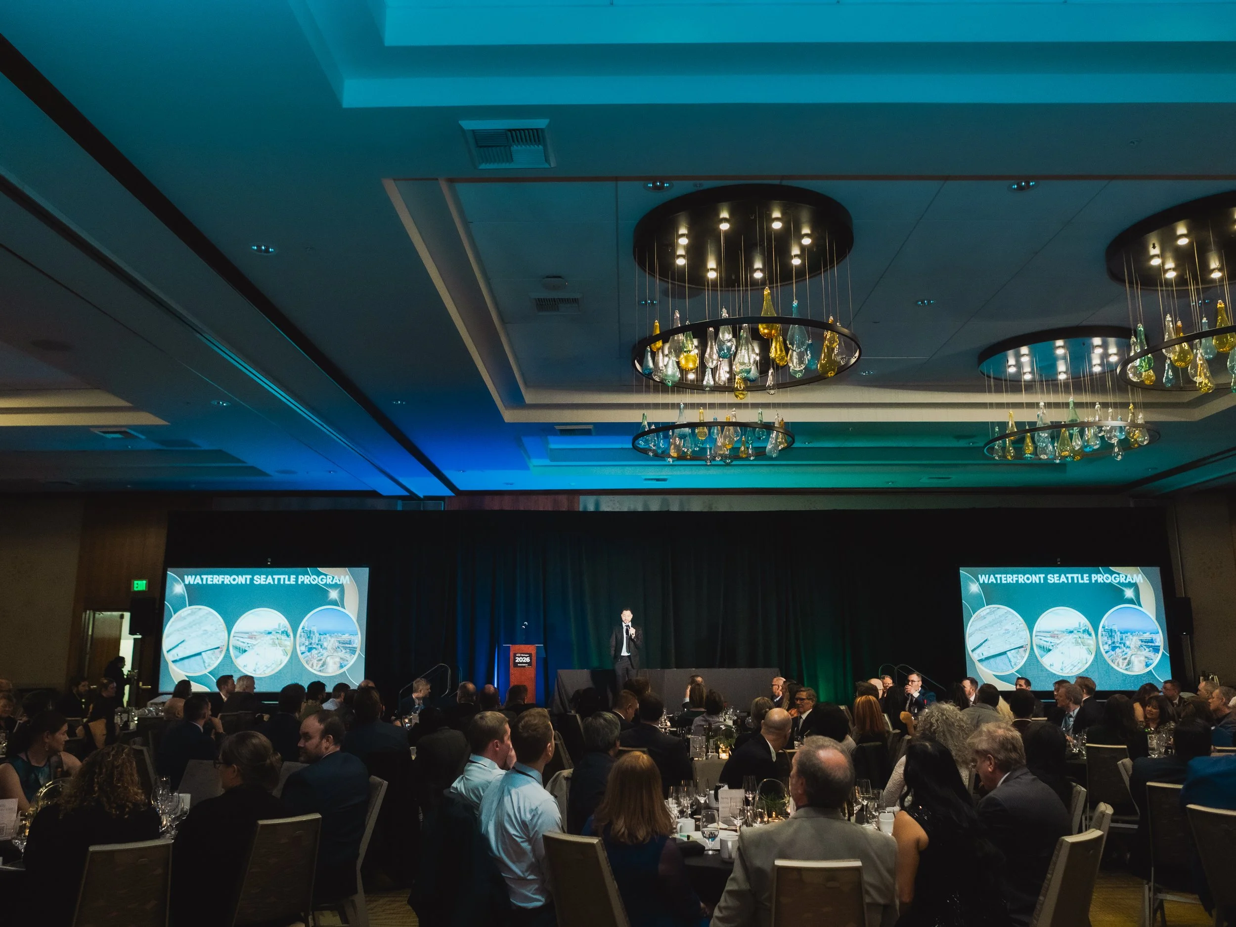 A speaker addresses a large audience seated at banquet tables during a formal conference at The Westin Bellevue. Presentation screens and modern chandeliers frame the ballroom.