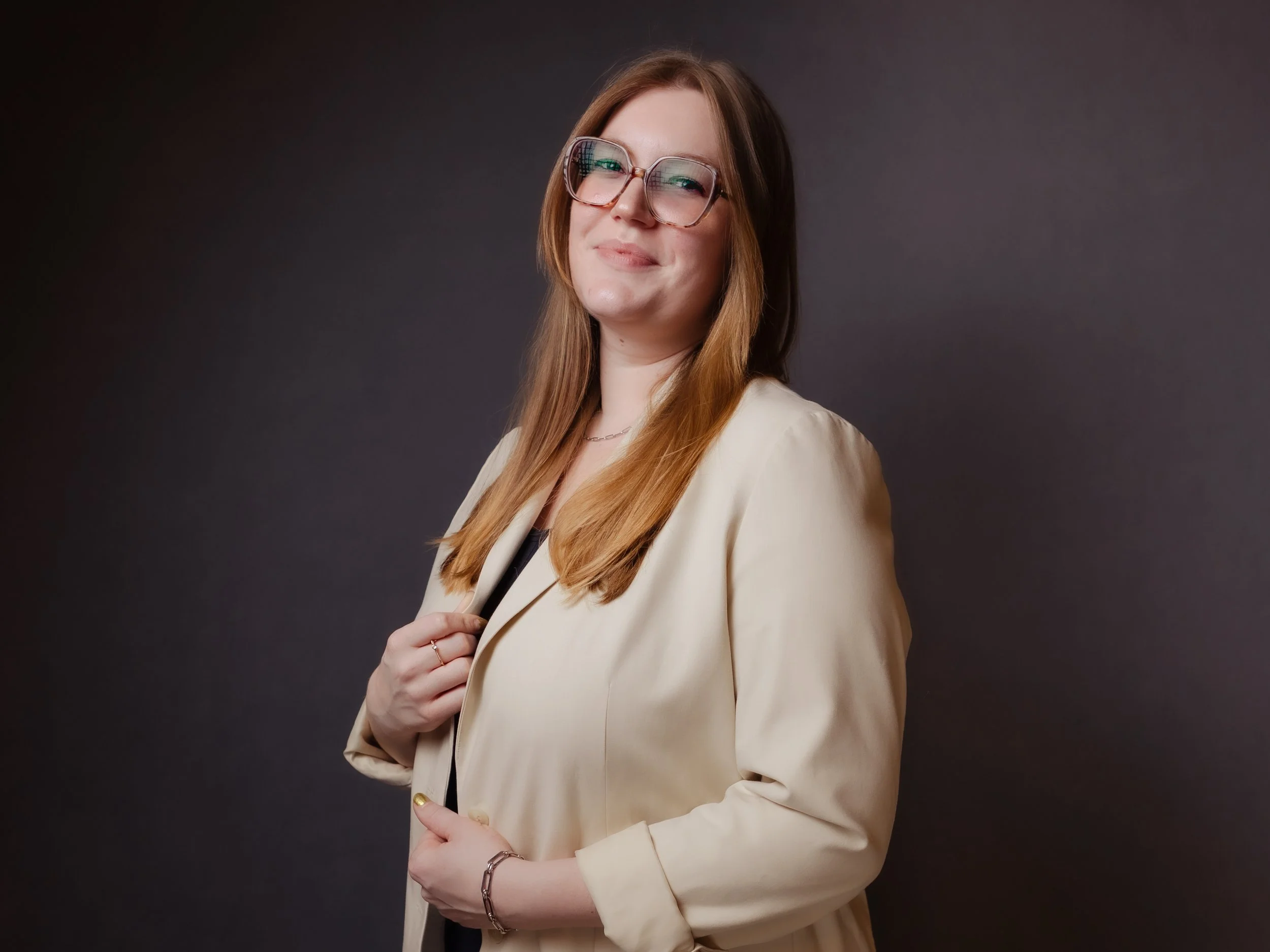 Professional headshot of Mairin McCurdy smiling softly while wearing glasses and a cream-colored blazer over a dark top. She stands against a dark studio background with her long light brown hair worn down.
