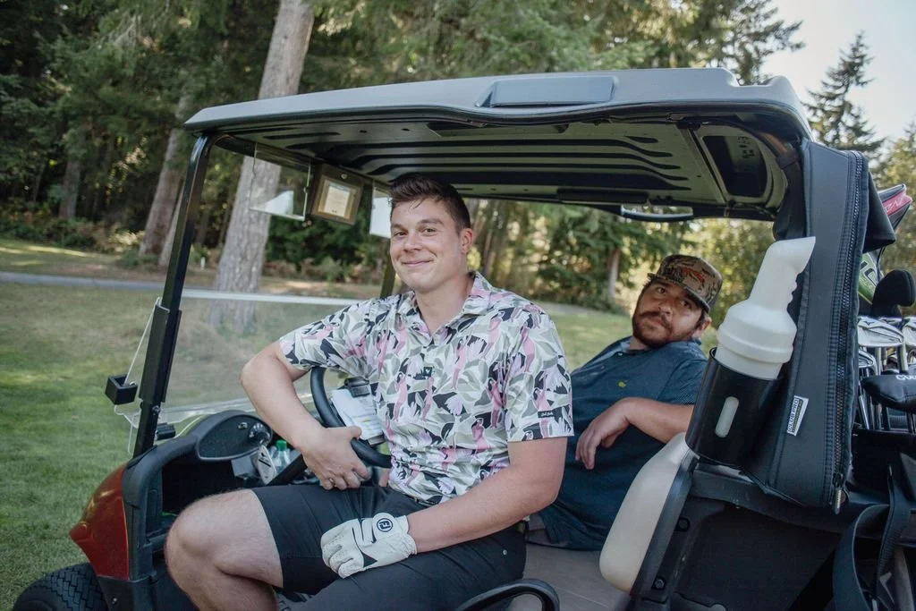 Two men sit in a golf cart at White Horse Golf Club in Kingston, Washington, smiling and relaxing near the green with trees in the background.