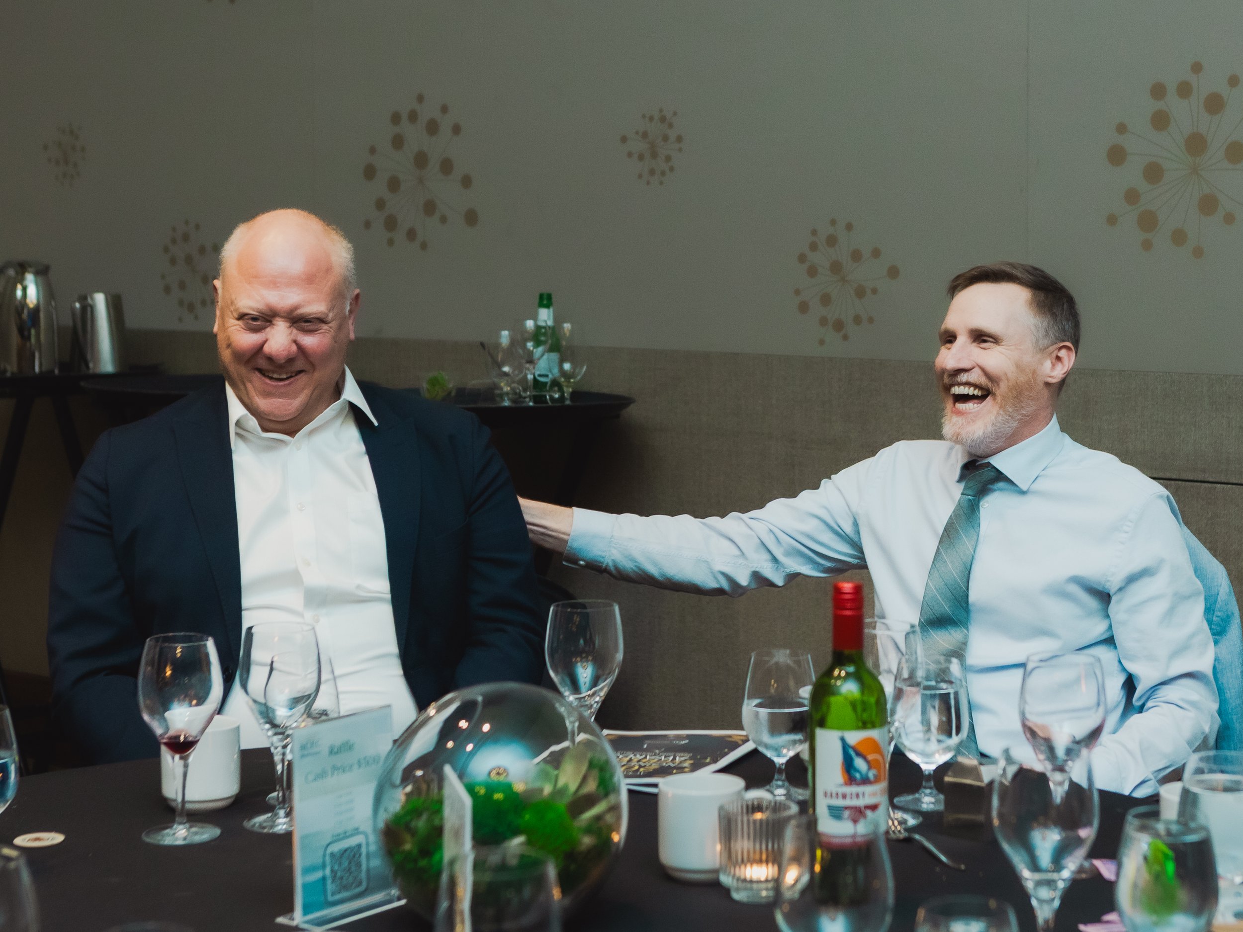 two men in business attire are laughing at a banquet table set with wine glasses and candles during a formal event