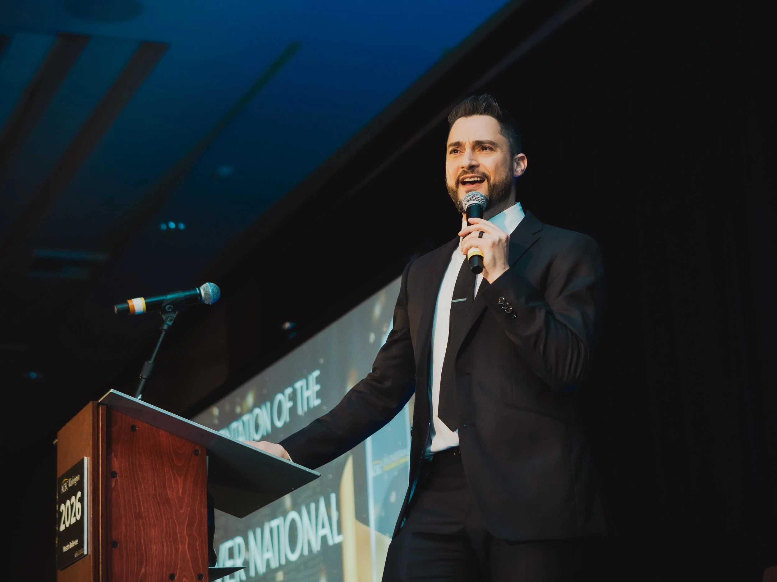 A man in a black suit and tie speaks into a microphone while standing at a podium during a formal presentation at The Westin Bellevue.