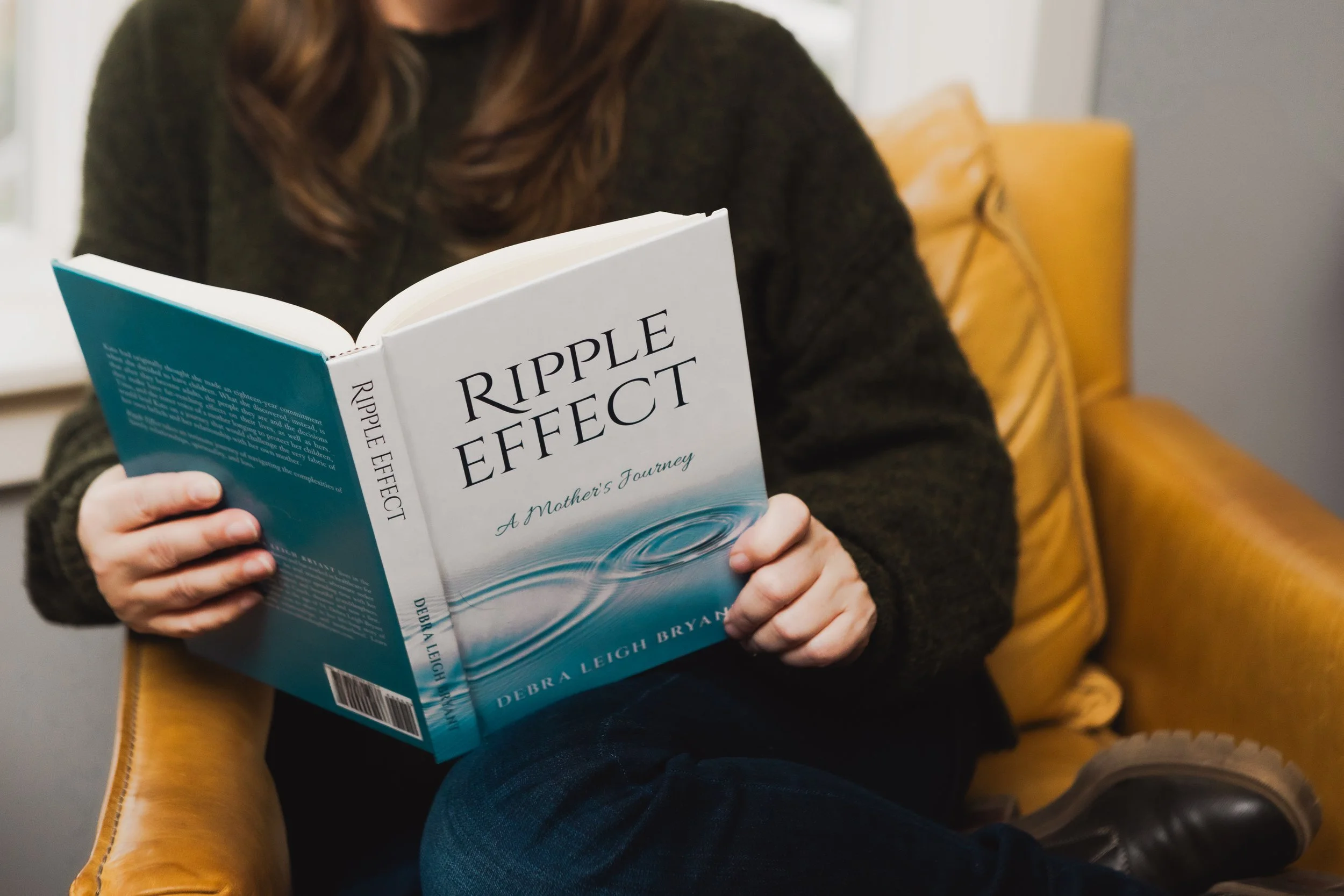 Close-up branding photograph of author Debra Bryant reading her book, Ripple Effect: A Mother's Journey, while seated in a leather chair.