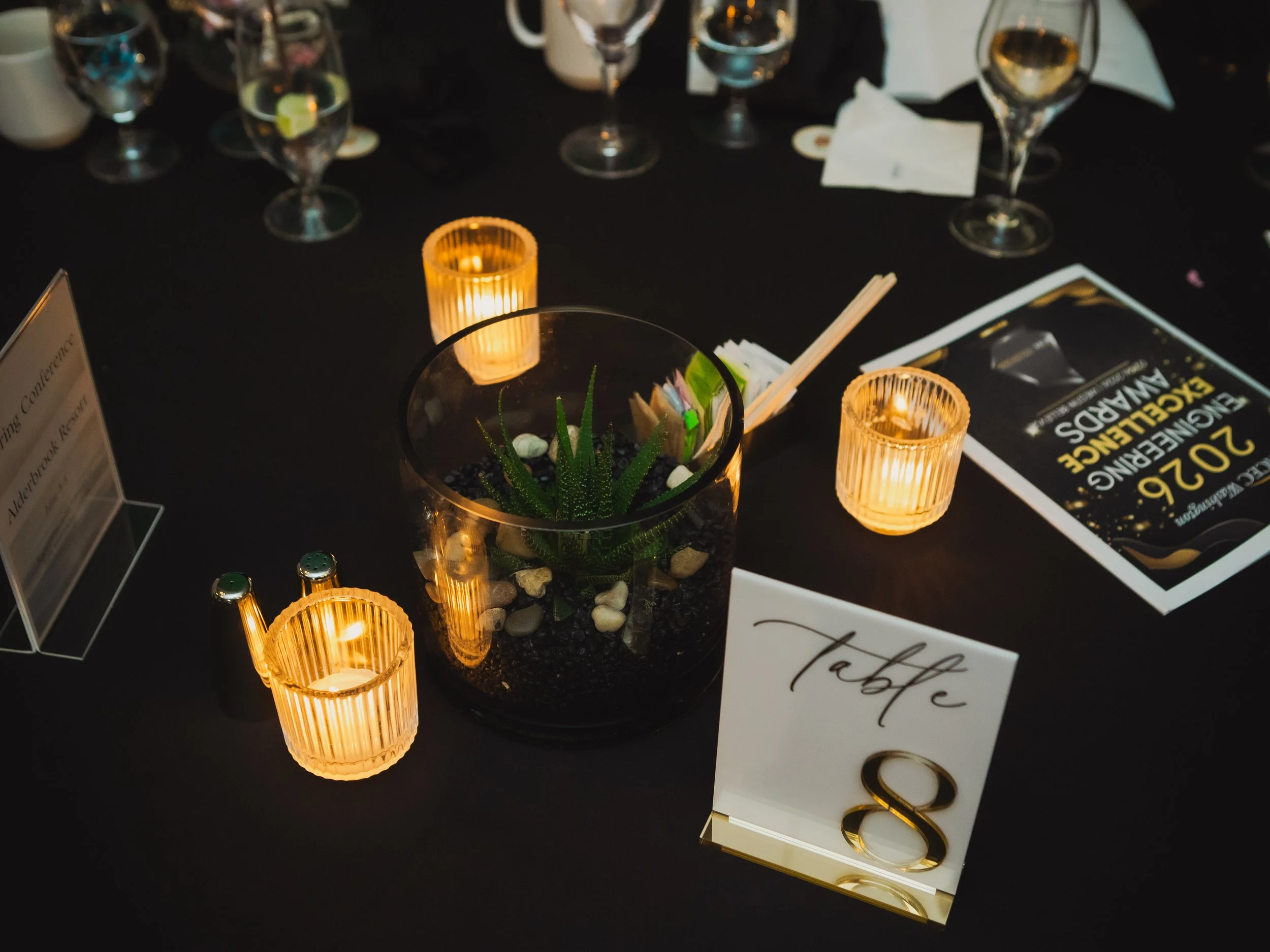 A banquet table centerpiece featuring candles, a glass vase with succulents, and a table number card rests beside a conference program and wine glasses.