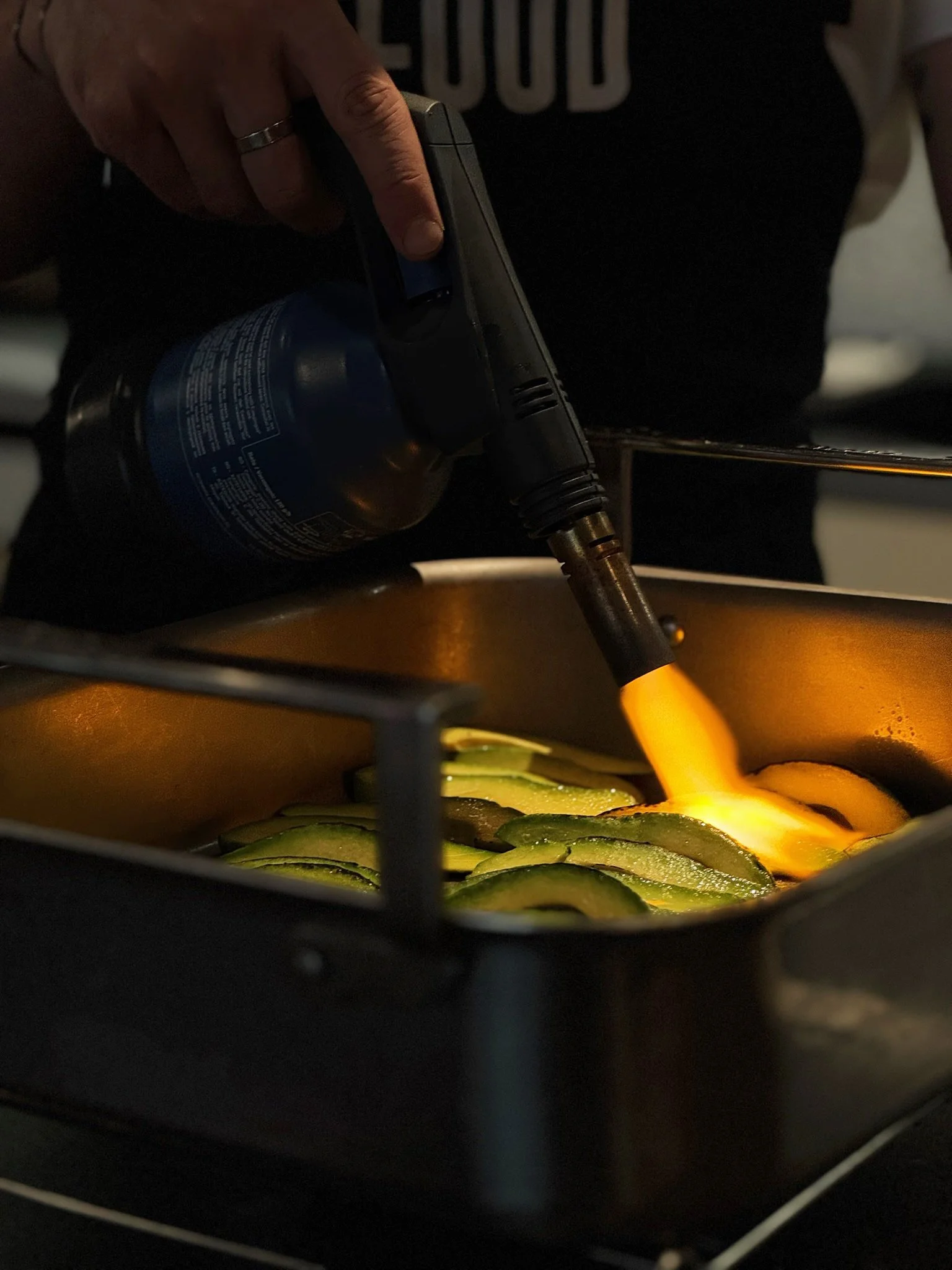 Une personne utilise un flamme torche pour faire griller ou caraméliser des légumes, probablement des courgettes, dans un plat de cuisson.