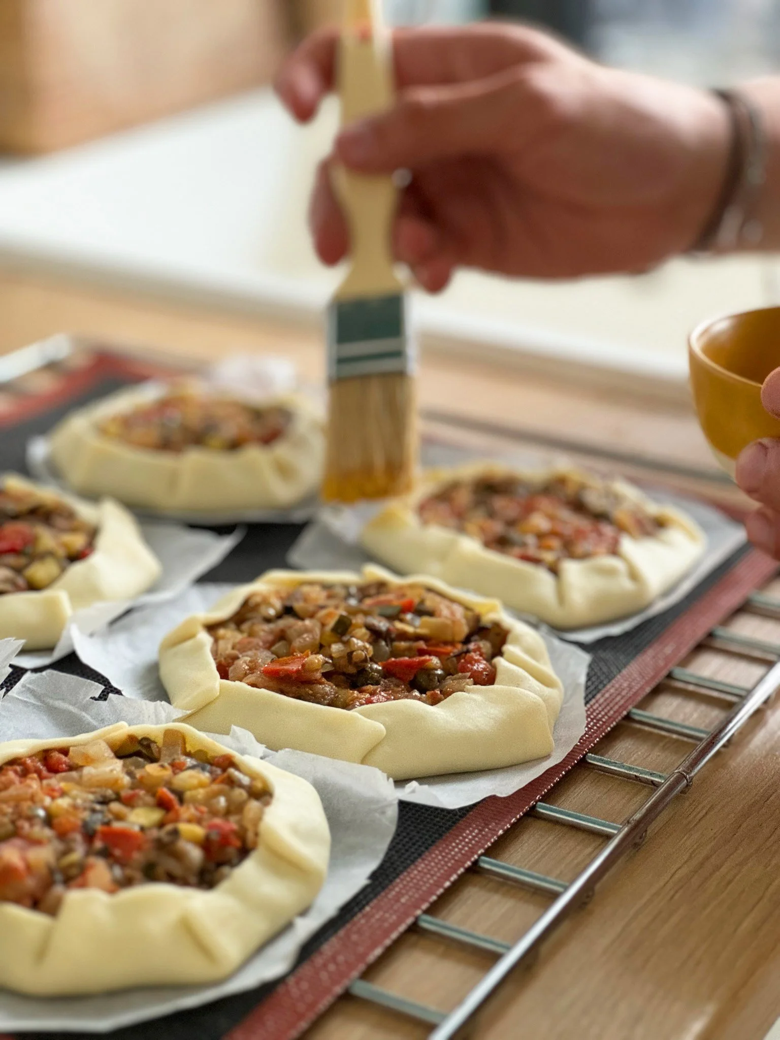 Main tenant une brosse à pâtisserie en train de badigeonner une pâte à tarte garnie de légumes et de viande, pour faire une pizza ou une tarte salée, sur une grille dans une cuisine.