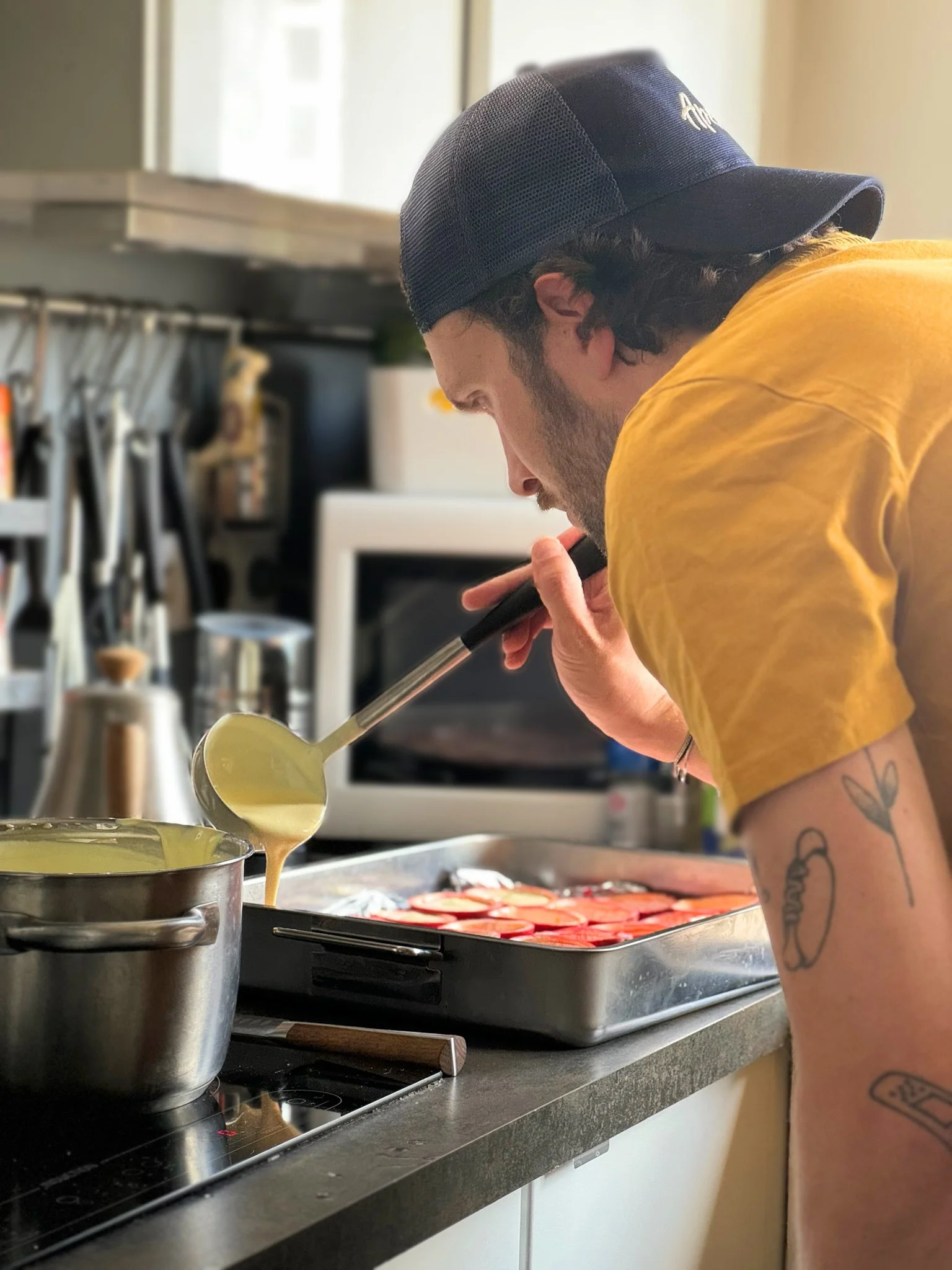 Un homme verse une sauce dans un plat de cuisine en utilisant une louche, dans une cuisine avec quelques ustensiles en arrière-plan.